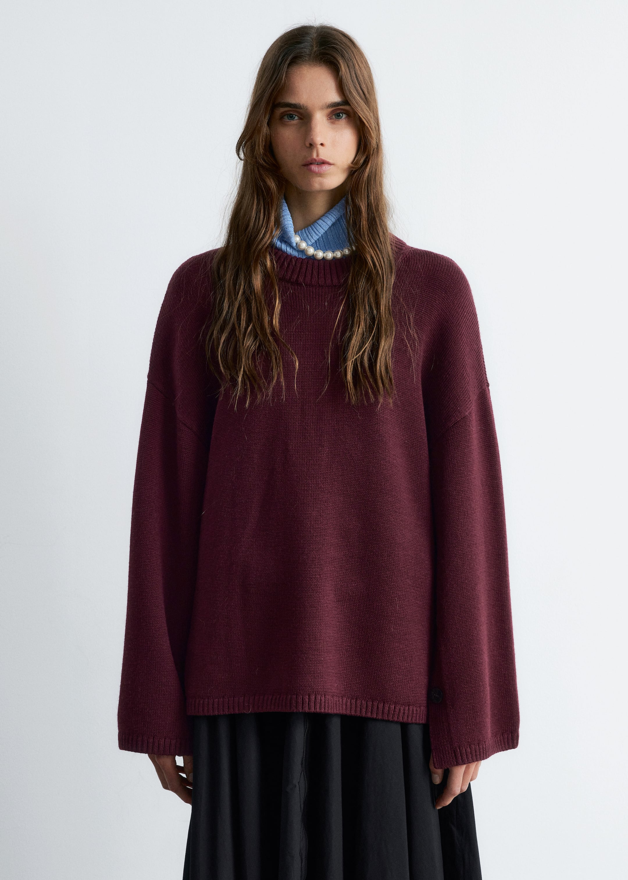 View larger image: A burgundy crew neck sweater is being presented from a front-facing angle, featuring a relaxed fit with ribbed cuffs and a ribbed hem. It is being layered over a light blue ribbed top and a pearl necklace, and paired with a black skirt.
