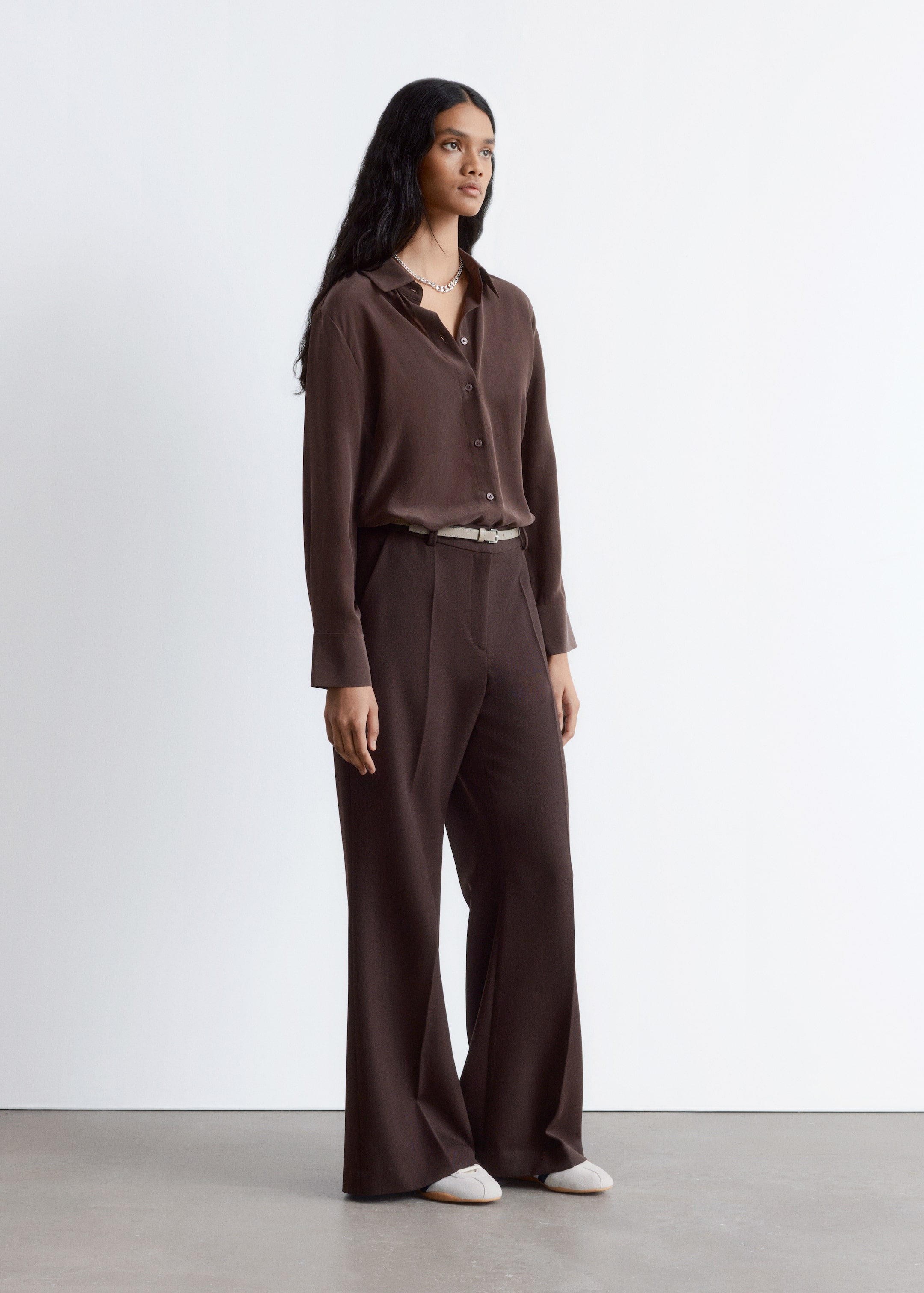 Visualizza immagine più grande: Una camicia di seta a maniche lunghe marrone scuro, con colletto a punta e polsini con bottoni, è indossata dal modello. La camicia è infilata in pantaloni a gamba larga marrone scuro e stretta con una cintura di colore chiaro, mentre una collana color argento è indossata sopra.