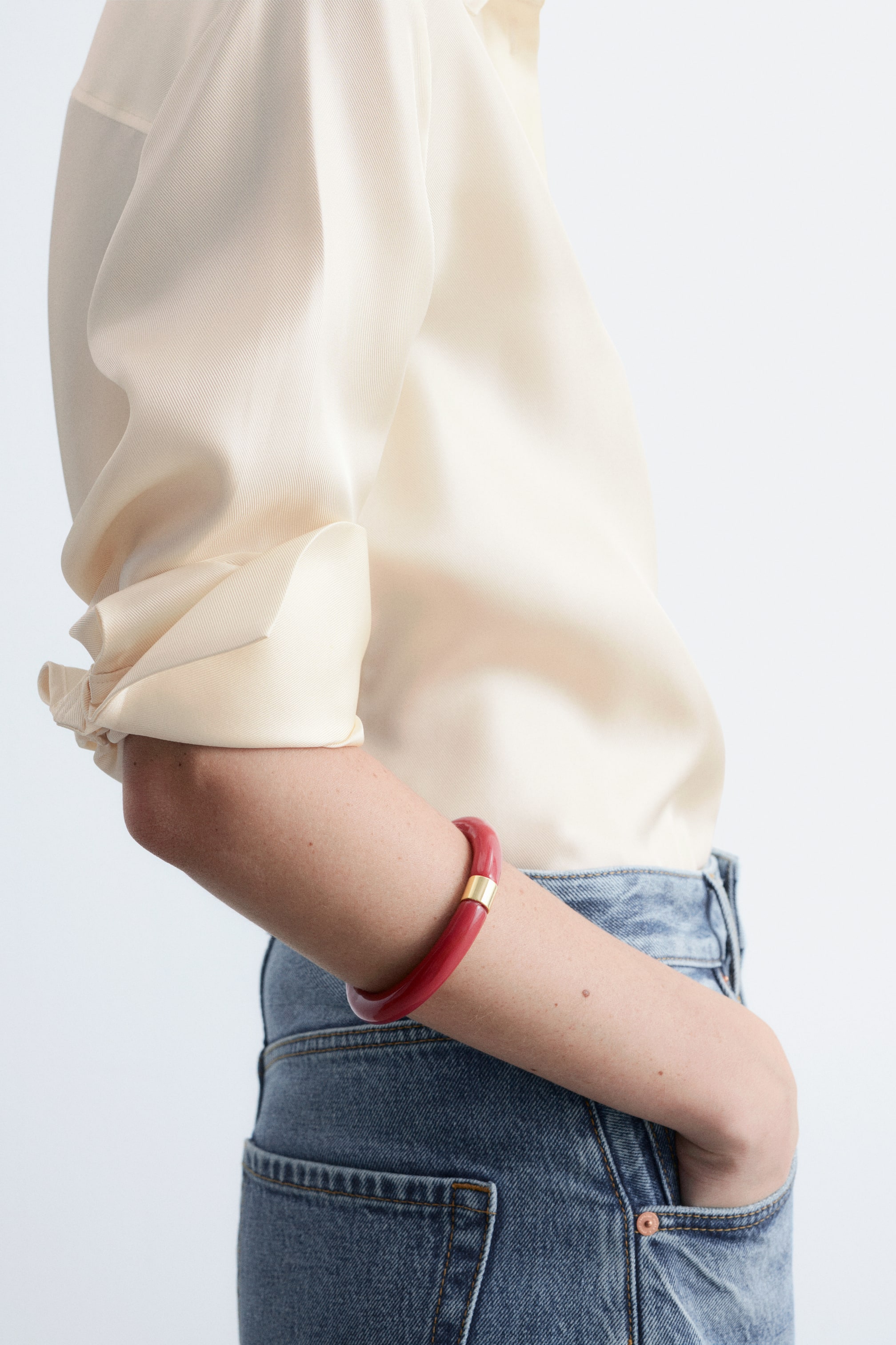 Grotere afbeelding bekijken: Een rode hars armband met een goudkleurig metalen detail wordt gedragen op de onderarm van het model, gestyled met een crèmekleurige blouse en een blauwe jeans.