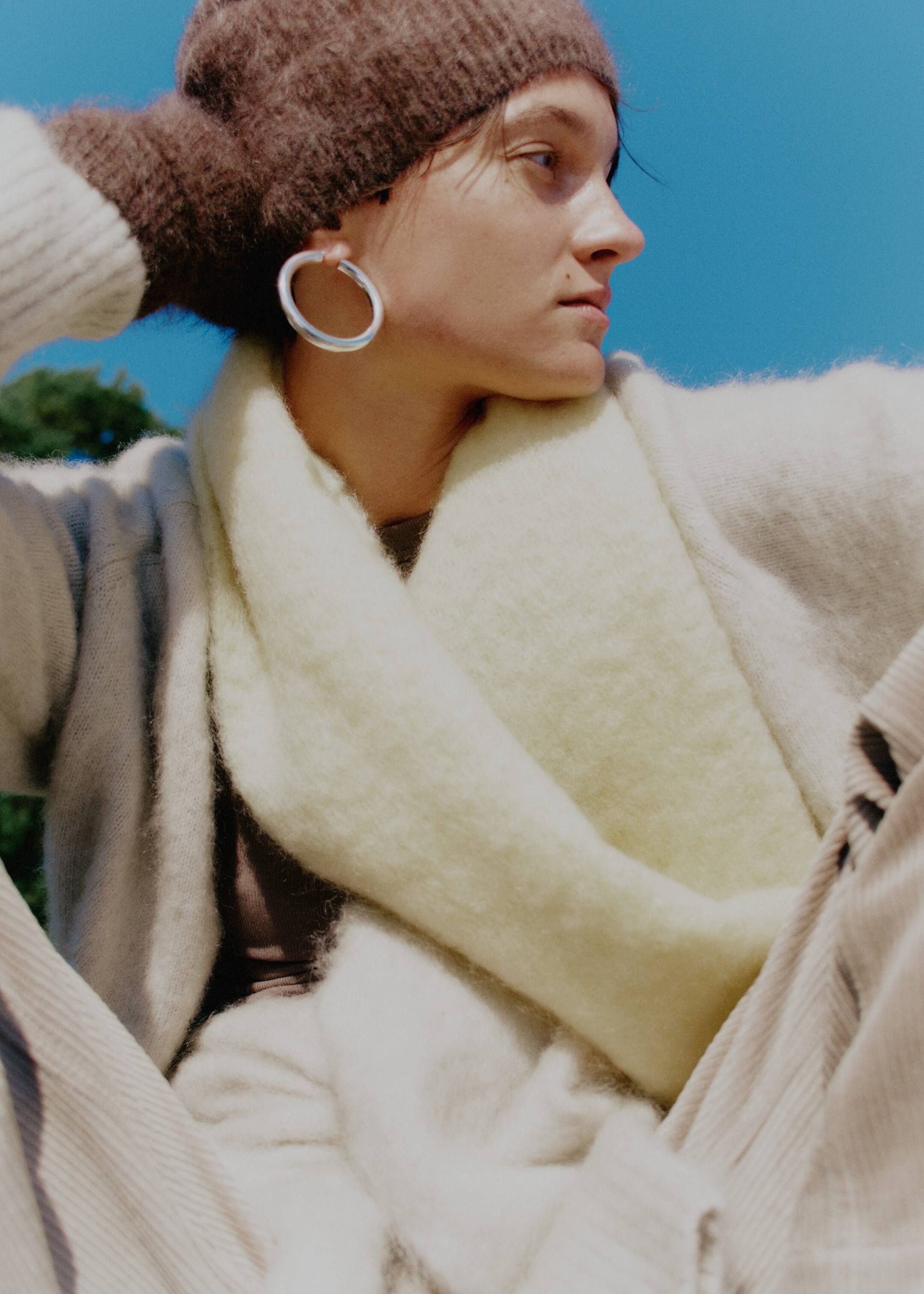 Größeres Bild anzeigen: Eine hellbeige, flauschige Strickjacke wird getragen, die über einen gerippten Bund verfügt und mit einem hellgelben, flauschigen Schal um den Hals drapiert geschichtet ist. Eine dunkelbraune, strukturierte Mütze bedeckt den Kopf, während ein dicker, silberfarbener Creolen-Ohrring leicht nach vorne geneigt am Ohr getragen wird.