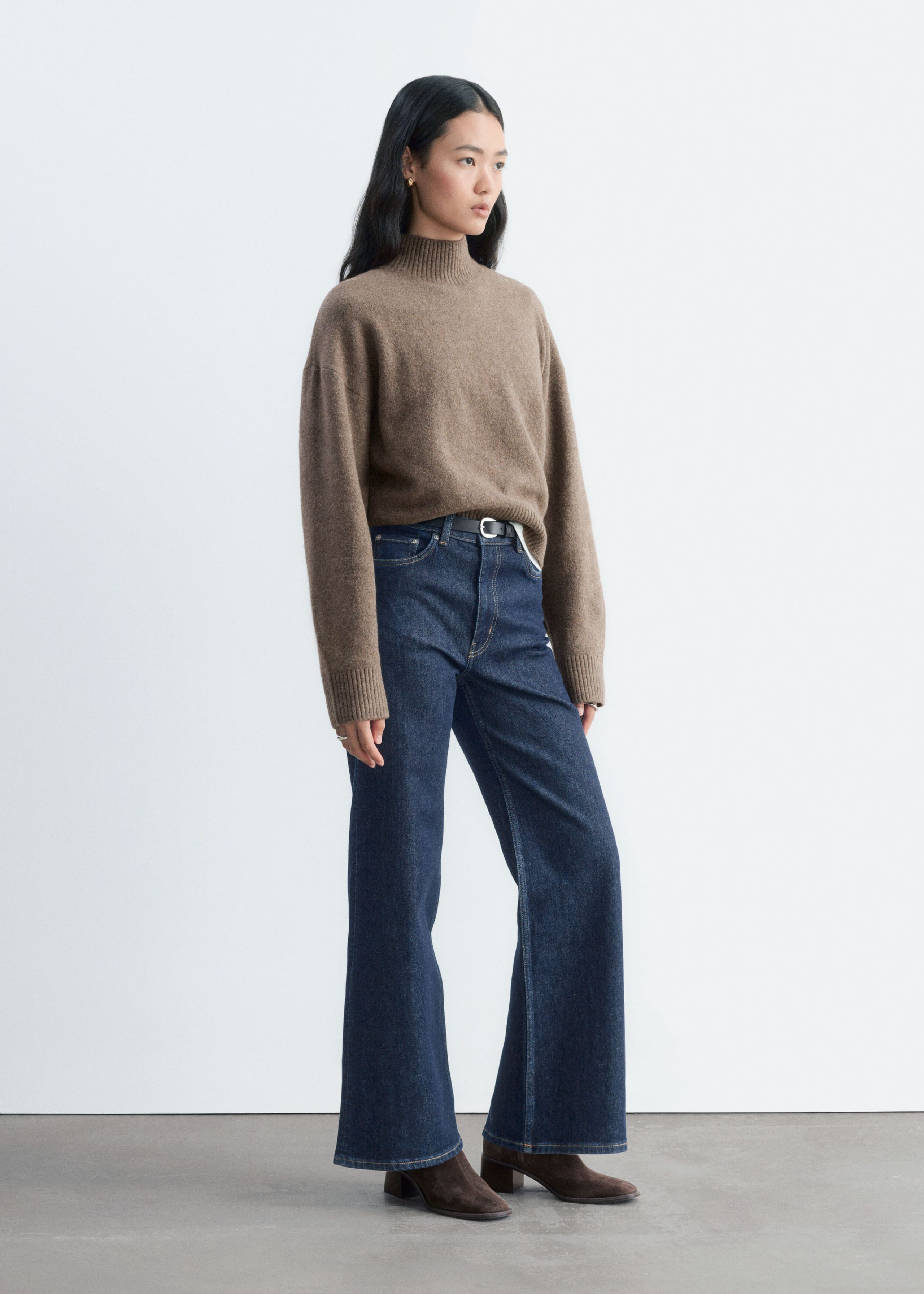 Een bruin gebreid trui met lange mouwen, voorzien van een geribde col, verlaagde schoudernaden en geribde boorden, wordt gedragen door het model. De losvallende trui is aan de voorkant ingestopt in een donkerblauwe wide-leg jeans.