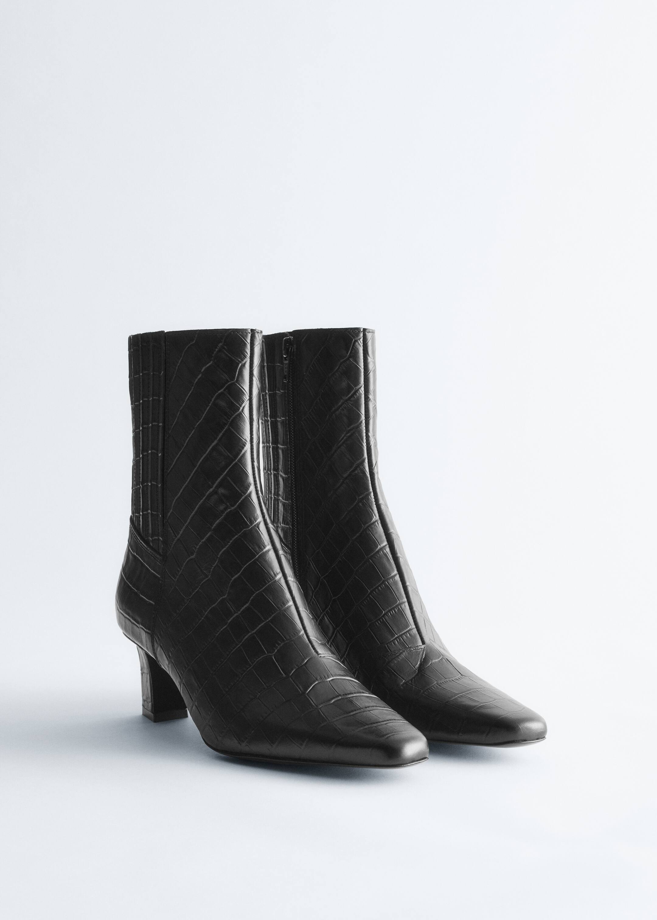 Ver imagen más grande: Un par de botines negros están de pie, mostrando su perfil frontal. Las botas presentan un patrón de cocodrilo grabado, punteras ligeramente cuadradas y una cremallera lateral en el lado interior de cada bota. Un panel acanalado es visible en la parte trasera de la caña, y las botas están asentadas sobre tacones bajos de bloque.