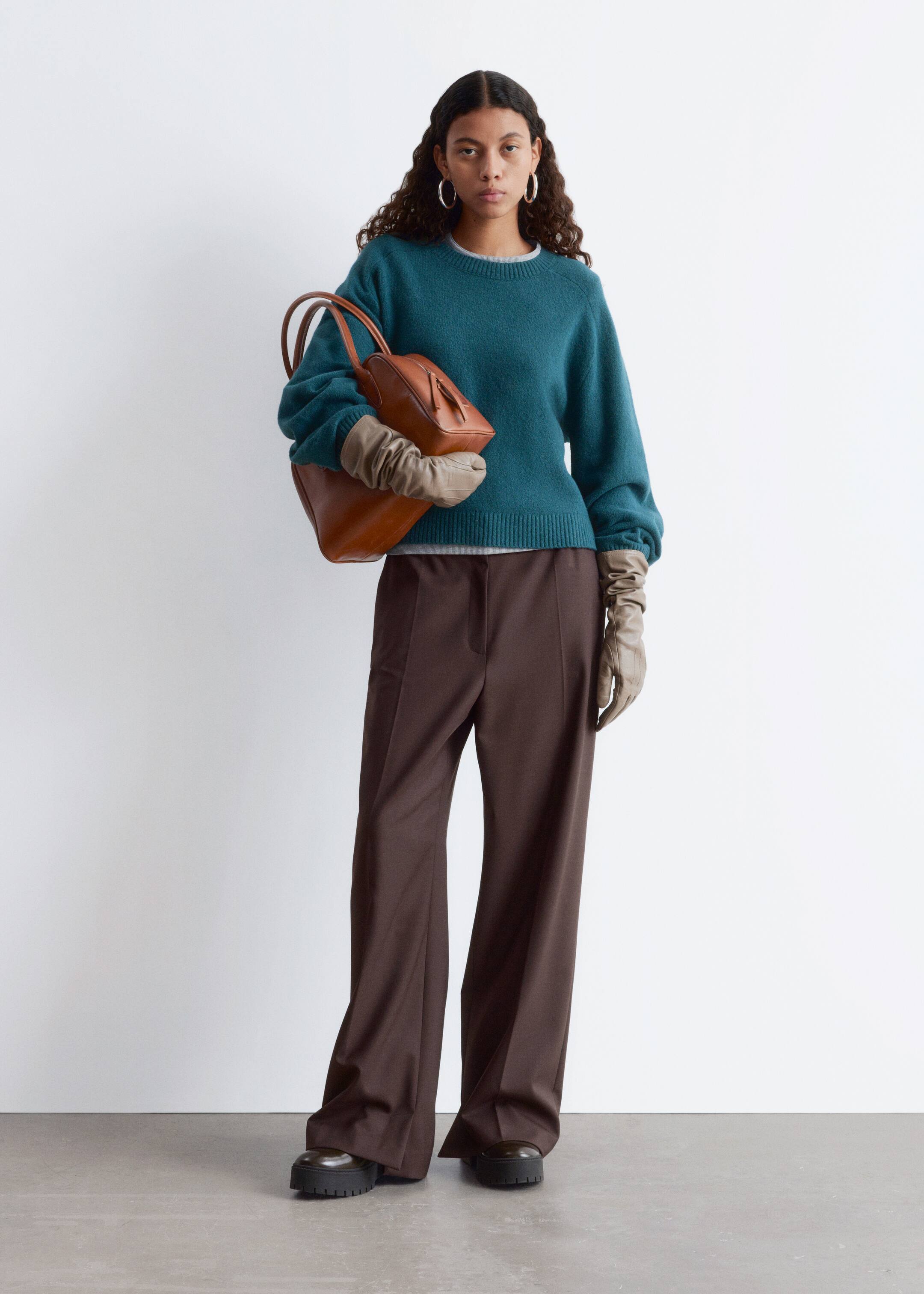 Grösseres Bild ansehen: Ein lockerer, gestrickter Pullover in Teal wird vom Model getragen, mit einem Rundhalsausschnitt, vollen Ärmeln mit gerafften Bündchen und gerippten Abschlüssen. Der Pullover ist über einem hellgrauen Oberteil geschichtet, und das Model hält eine braune Handtasche, während sie lange beige Handschuhe trägt.