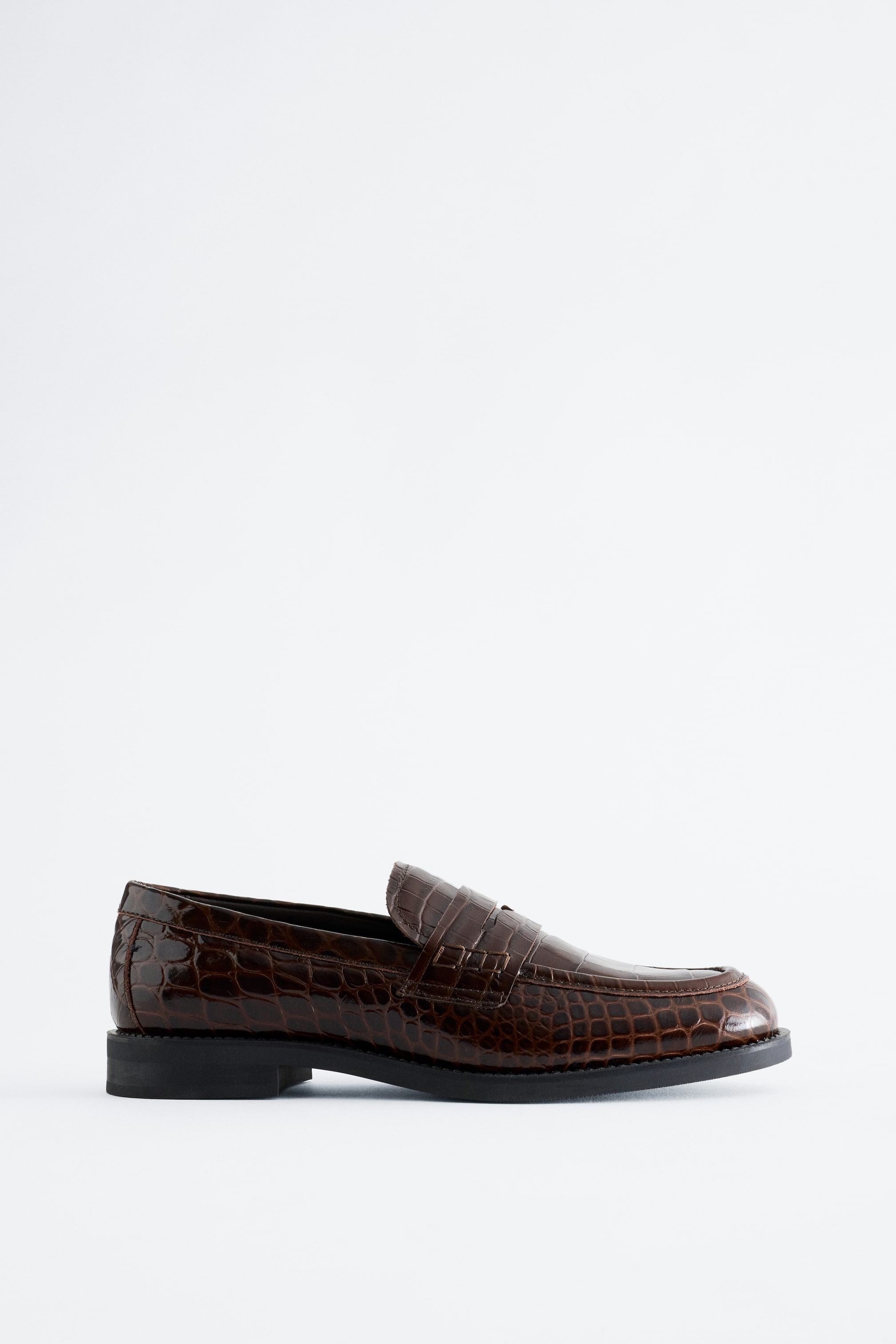 Visa större bild: En klassisk penny loafer i högblankt, krokodilpräglat brunt läder visas i sidoprofil. Skon har toppsticksdetaljer runt tån, en traditionell pennyrem över vristpartiet och en tjock svart gummisula.
