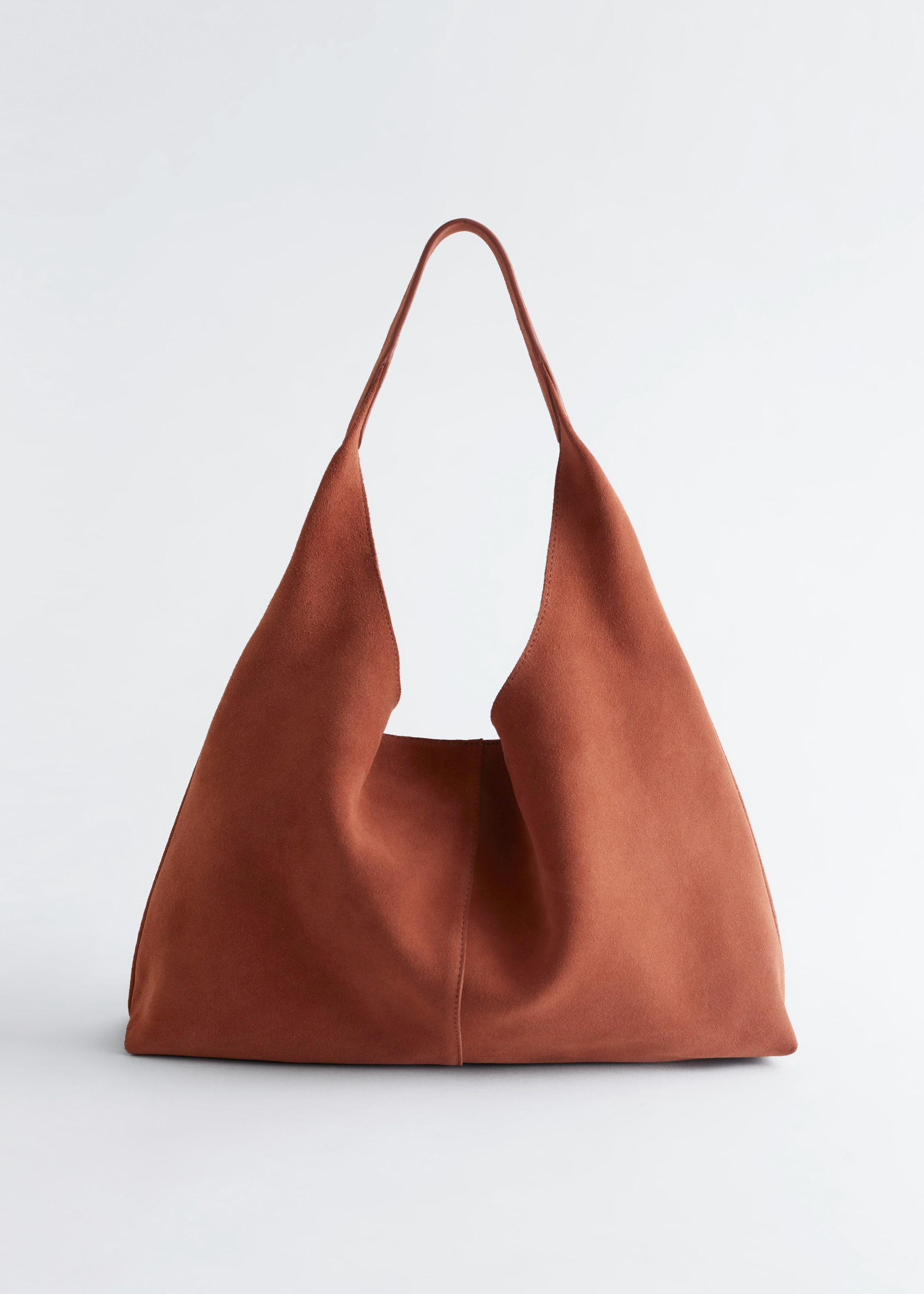 Ver imagem maior: Um saco de tote castanho-avermelhado está apresentando a sua vista FRONTAL, feito de camurça lisa e mate. Está a apresentar uma costura vertical decorativa que desce pelo centro da frente, com uma única alça larga que se estende para cima.