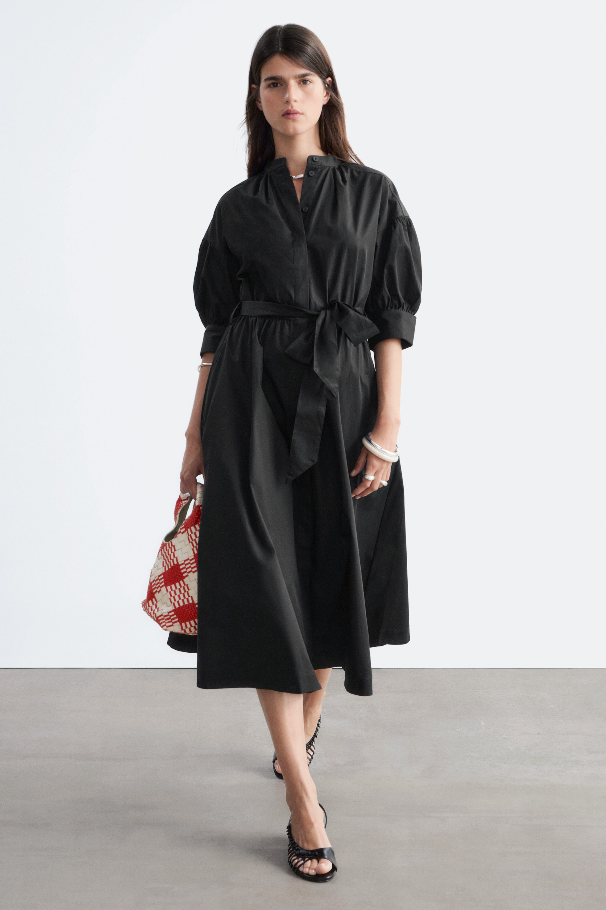 Se større billede: En sort midikjole med en båndkrave og en delvis knaplukning. Den er designet med voluminøse trekvartærmer, der er samlet ved manchetterne, og et bindebælte, der fremhæver taljen på dens flydende midinederdel.
