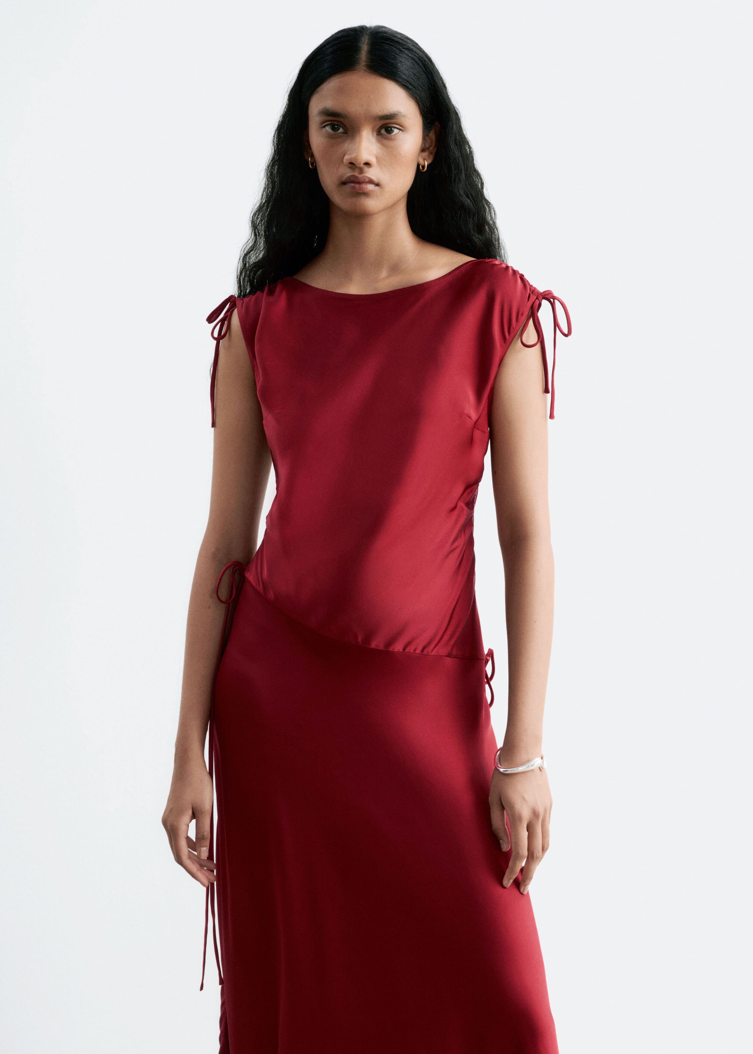 The model is wearing a dark red satin dress featuring drawstring details at the shoulders and a boat neckline. The dress is designed with an asymmetric dropped waist, showcasing gathered fabric and side ties.