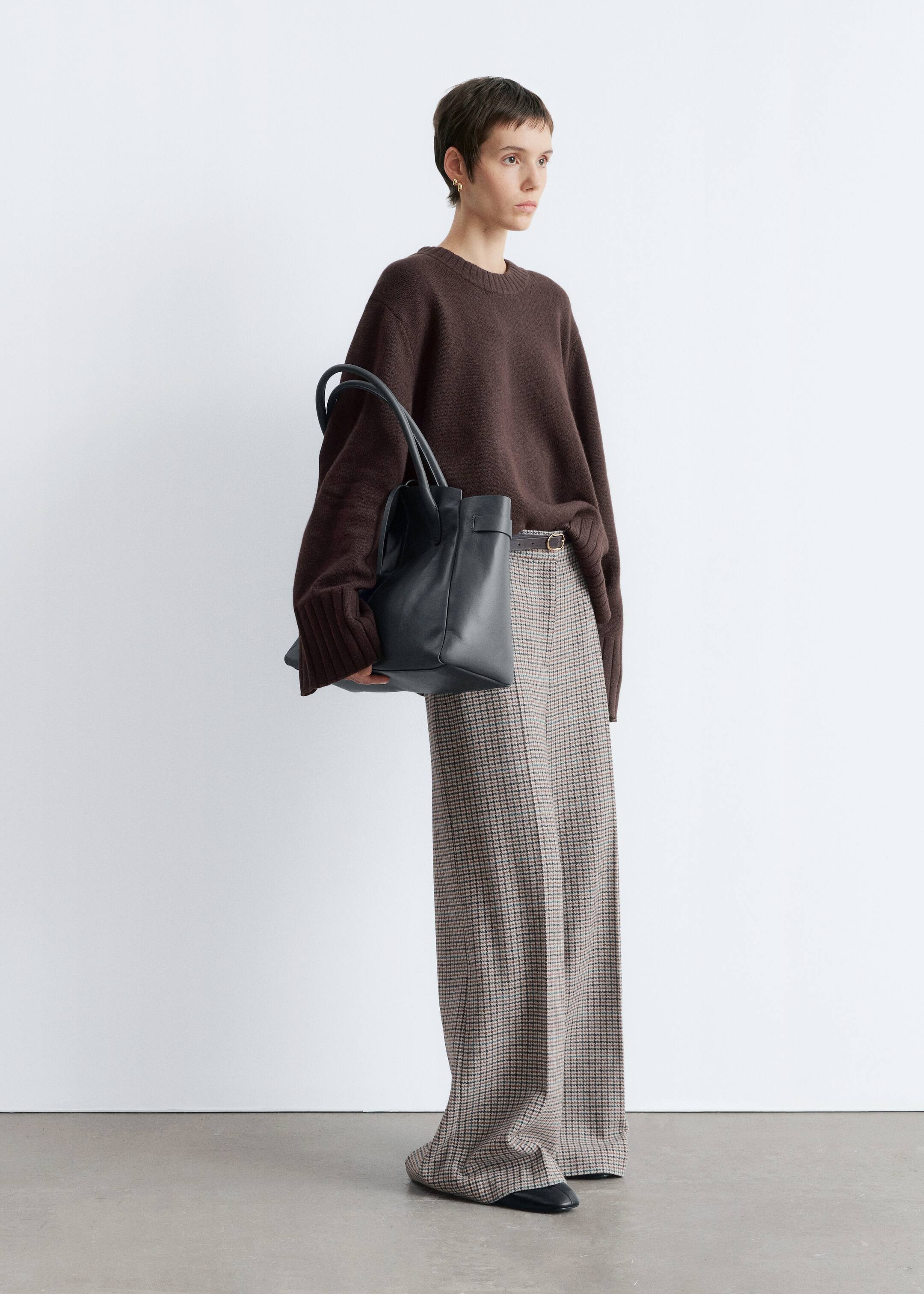 View larger image: A relaxed-fit, dark brown crew neck sweater with ribbed cuffs is being styled with wide-leg plaid trousers, which are being cinched by a thin dark belt. A black structured tote bag with two top handles is being held, and black pointed-toe flat shoes are completing the ensemble.