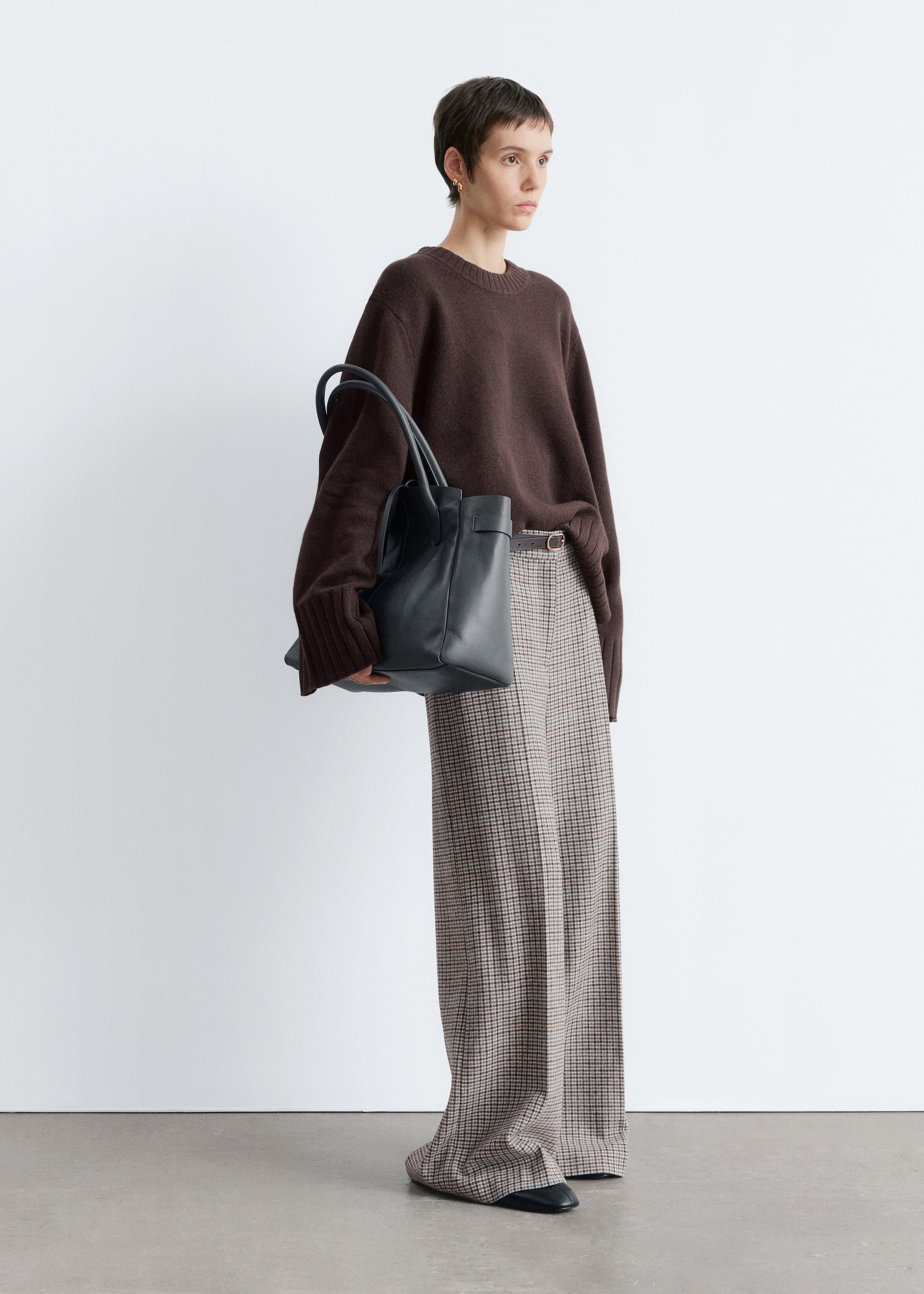Agrandir l'image: Un pull à col rond marron foncé à coupe décontractée avec des poignets côtelés est associé à un pantalon à carreaux à jambes larges, qui est ceinturé par une fine ceinture sombre. Un sac fourre-tout structuré noir avec deux poignées supérieures est tenu, et des chaussures plates à bout pointu noires complètent l'ensemble.