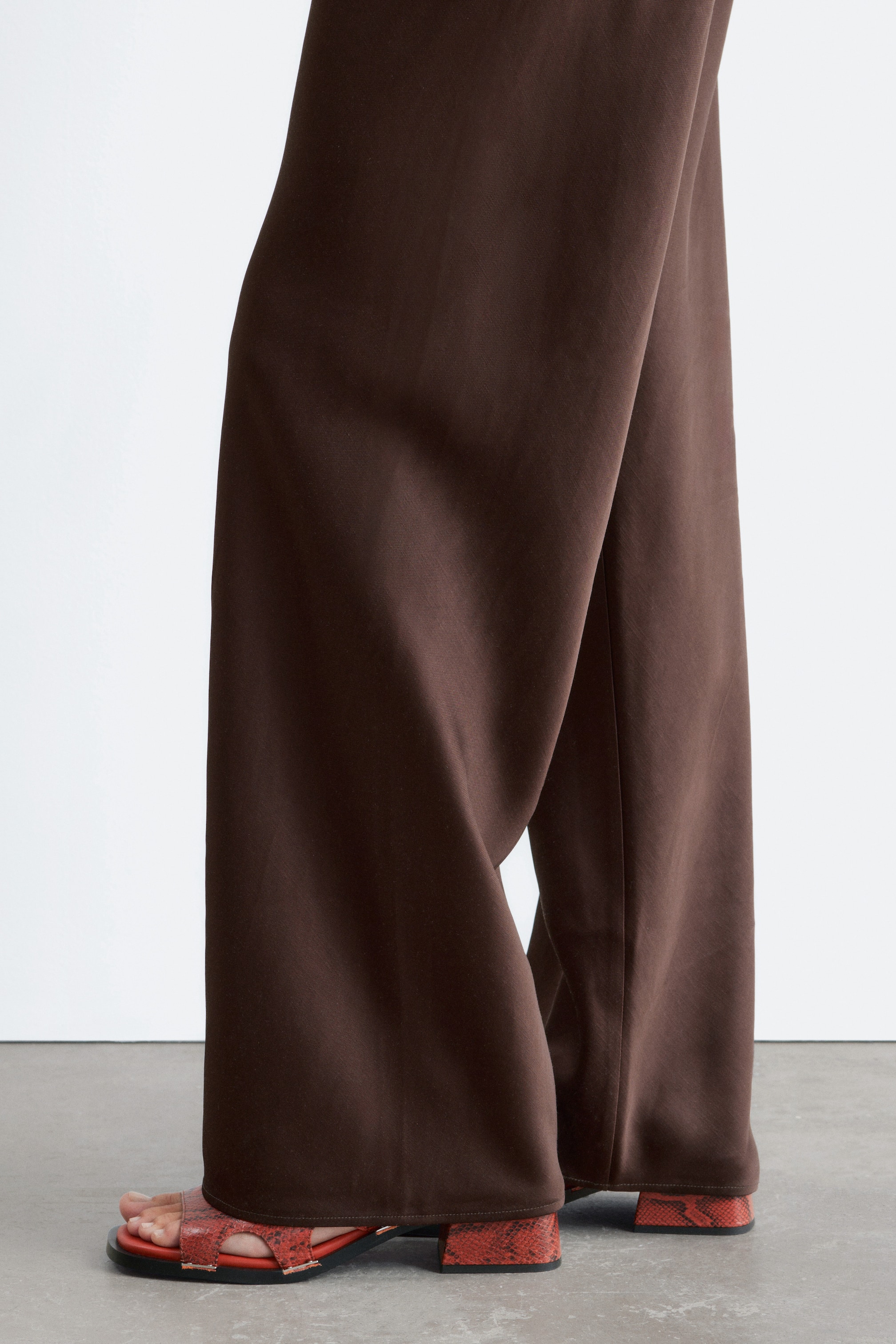 Zobrazit větší obrázek: Hnědé kalhoty s širokými nohavicemi splývají dolů a zakrývají vrchní část sandálů. Kalhoty se lehce hromadí u kotníků, což zdůrazňuje jejich plnou délku a jemný splývavý střih.