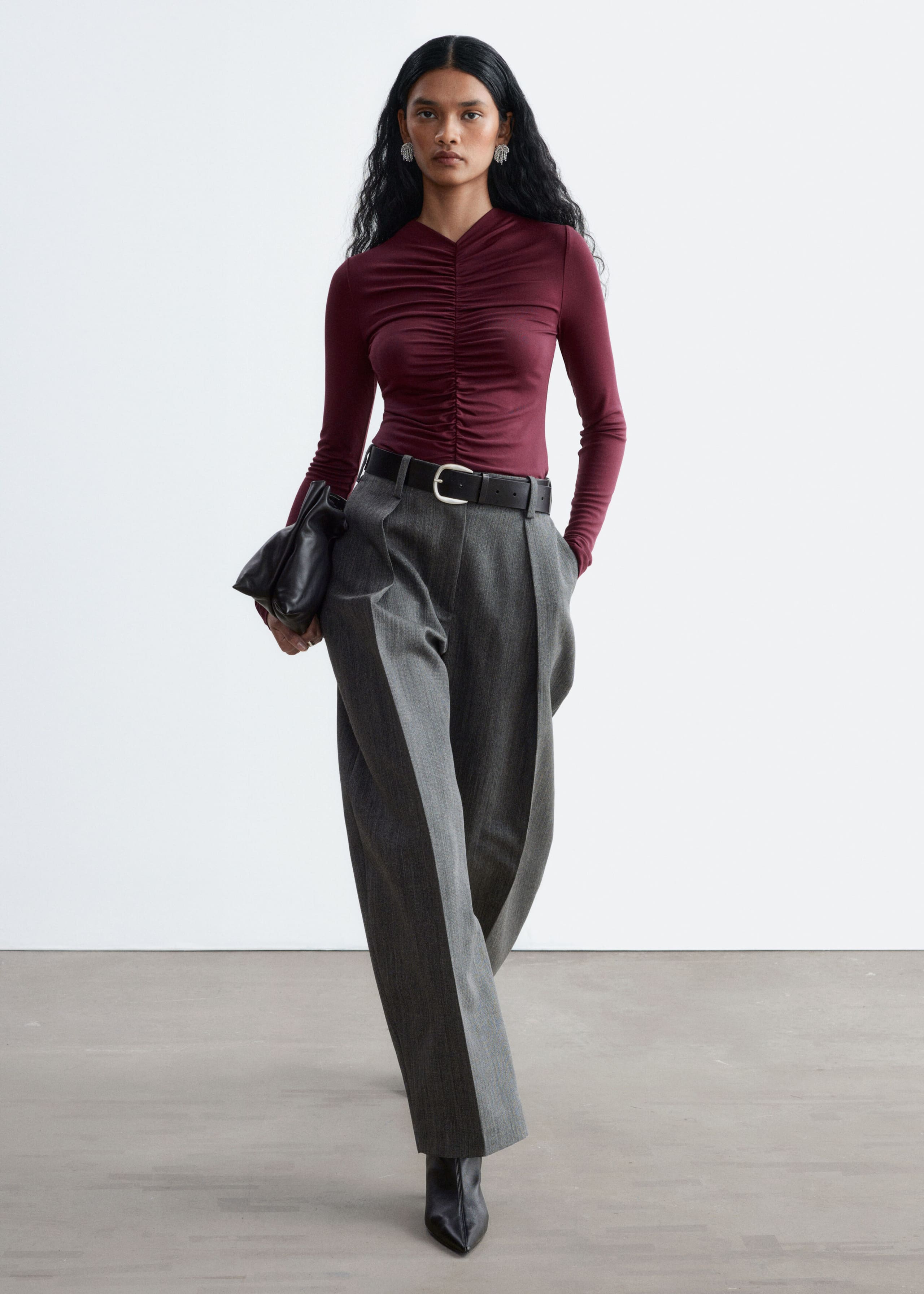 A long-sleeved burgundy top featuring detailed gathering along the center front seam is being worn by the model. The fitted top is tucked into high-waisted gray trousers with a black belt, while the model is holding a black clutch bag.