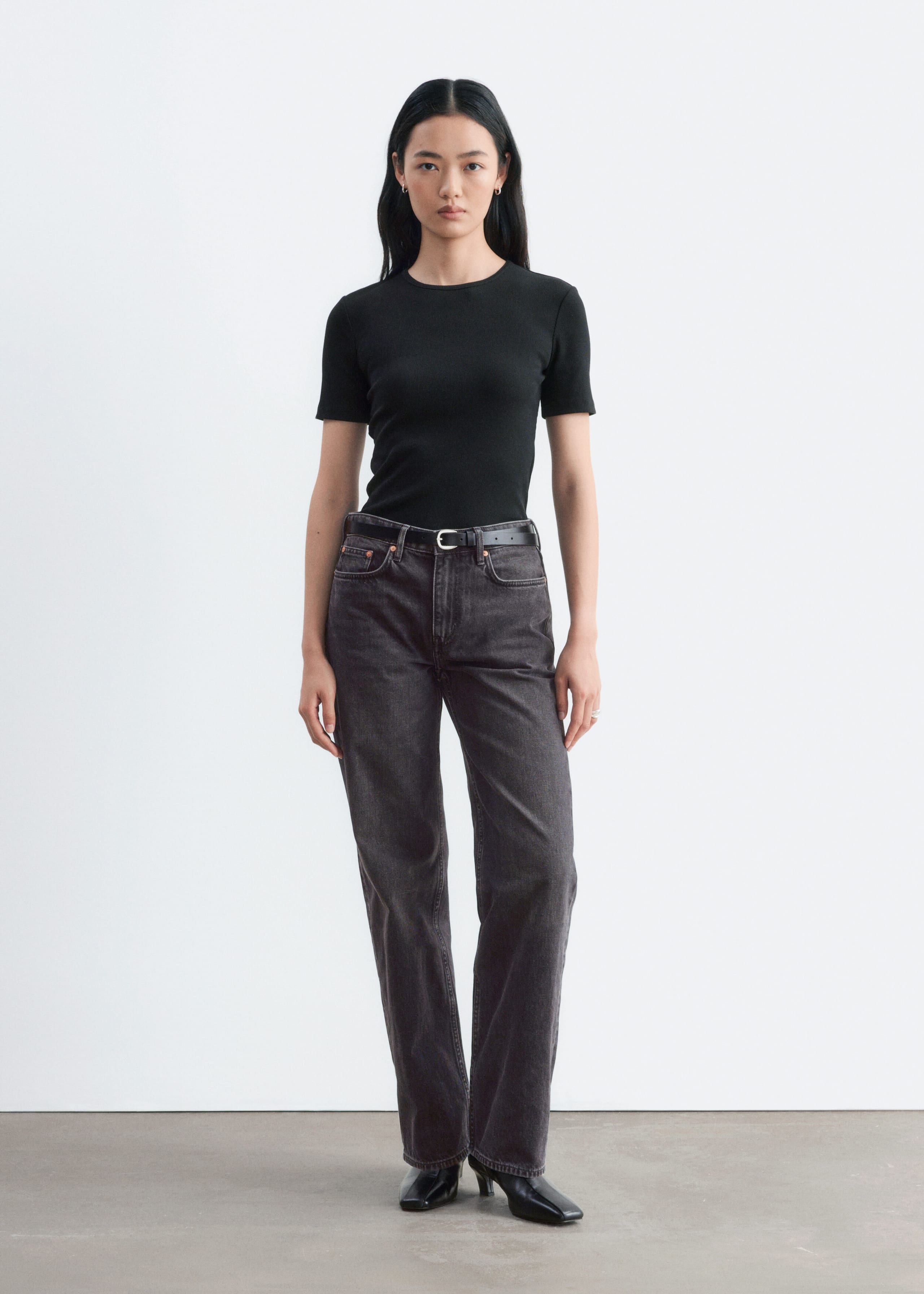 A black, slim-fit ribbed T-shirt with a crew neck and short sleeves is being worn by the model. The T-shirt is tucked into dark grey jeans styled with a black belt.