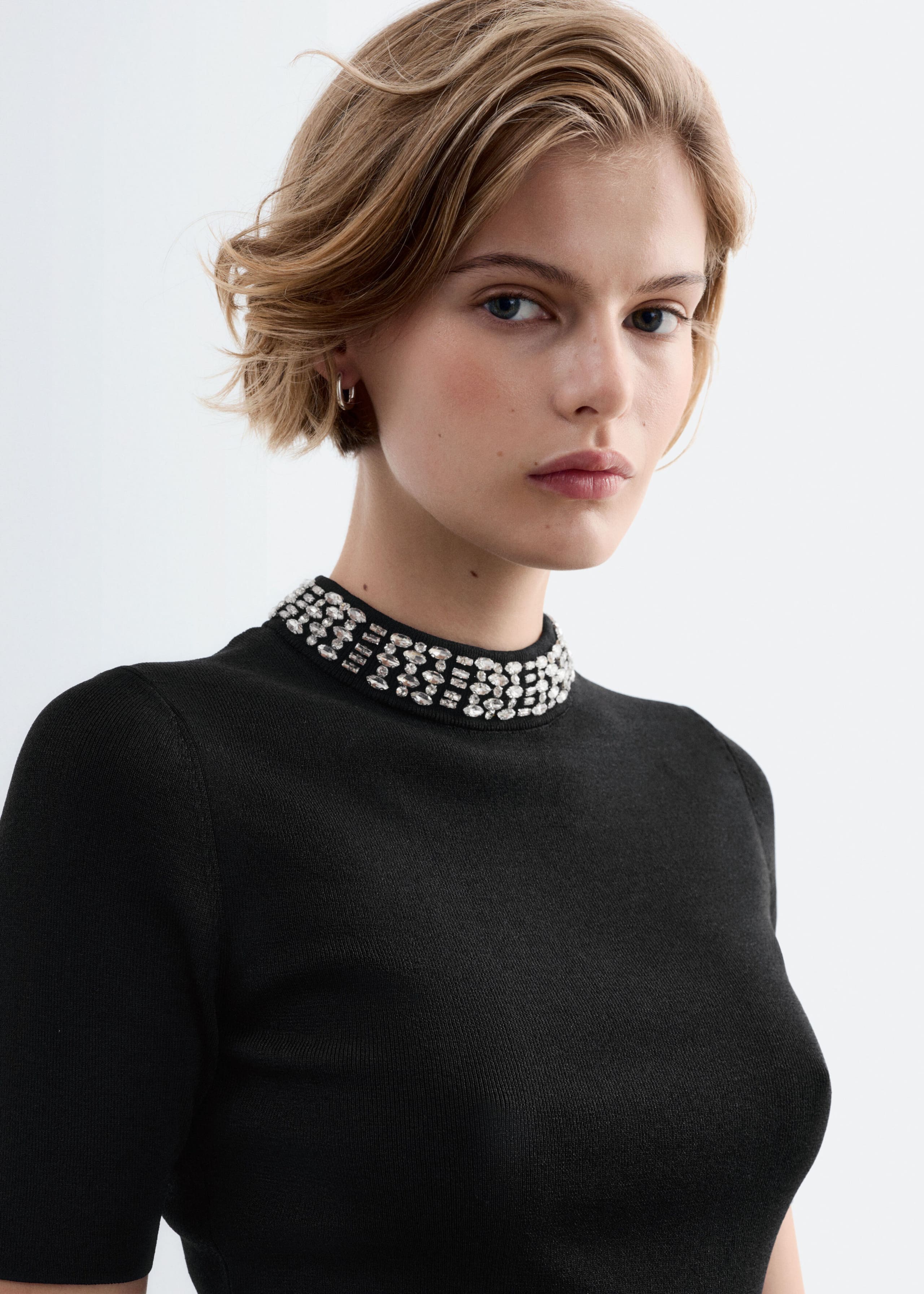 The model is wearing a black fitted knit top with a mock neck accented by rows of silver rhinestone beads.