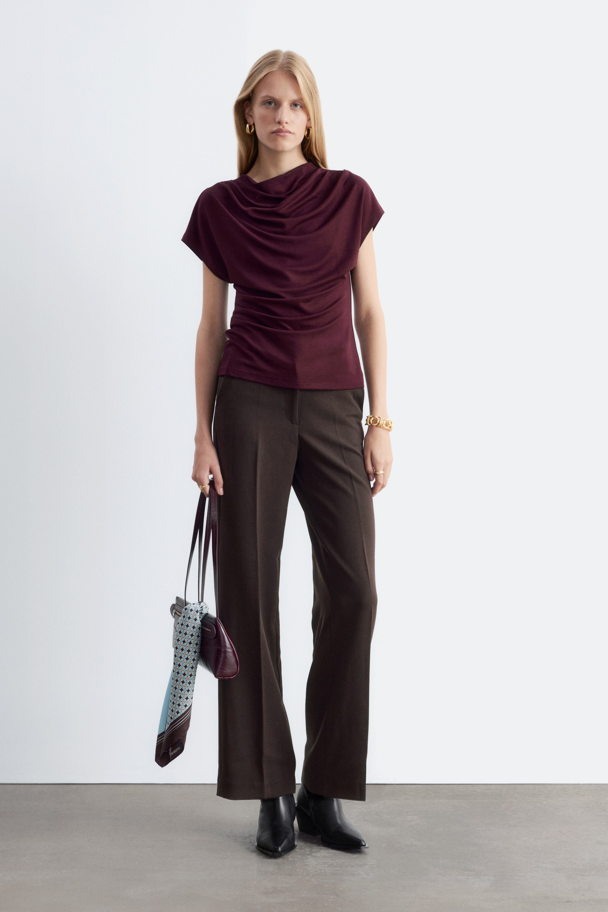 Größeres Bild ansehen: Dunkelbraune Straight-Leg-Hose mit markanten Bügelfalten vorne, getragen von dem Model, das frontal steht. Die Hose ist kombiniert mit einem drapierten, ärmellosen Oberteil in Burgunderrot, schwarzen Stiefeletten mit spitzer Spitze und einer kleinen Handtasche, die in einer Hand gehalten wird.