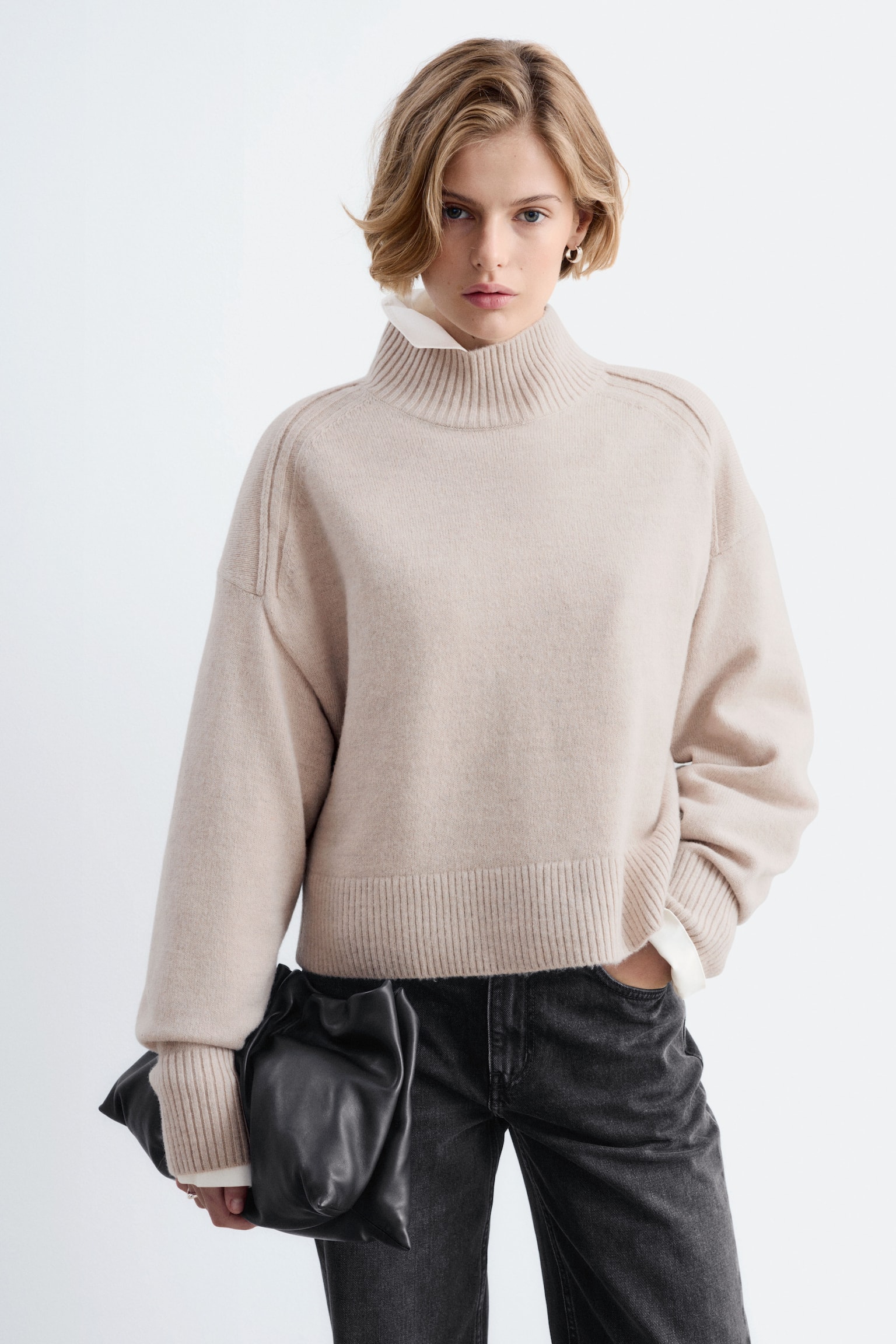 Un maglione beige con collo a lupetto Ã¨ ripreso frontalmente, mettendo in risalto una silhouette rilassata con cuciture delle spalle in avanti. Presenta un colletto a costine, un ampio bordo a costine e maniche lunghe con polsini allungati a costine. - 1