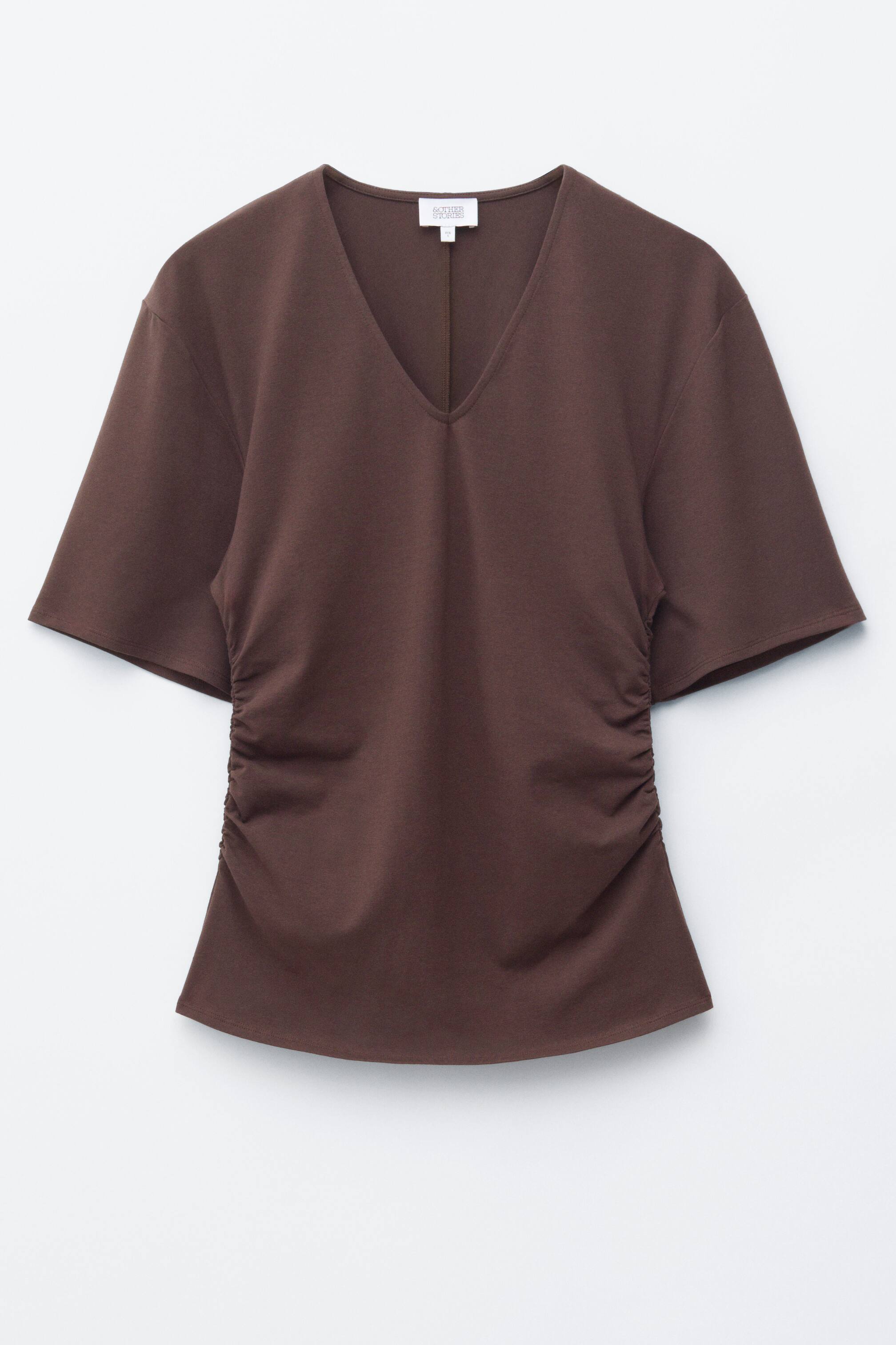 Ver imagem maior: Uma T-shirt castanha está voltada para a frente, apresentando um decote em V e mangas curtas generosas. Pregas são visíveis ao longo das costuras laterais, criando um efeito franzido.