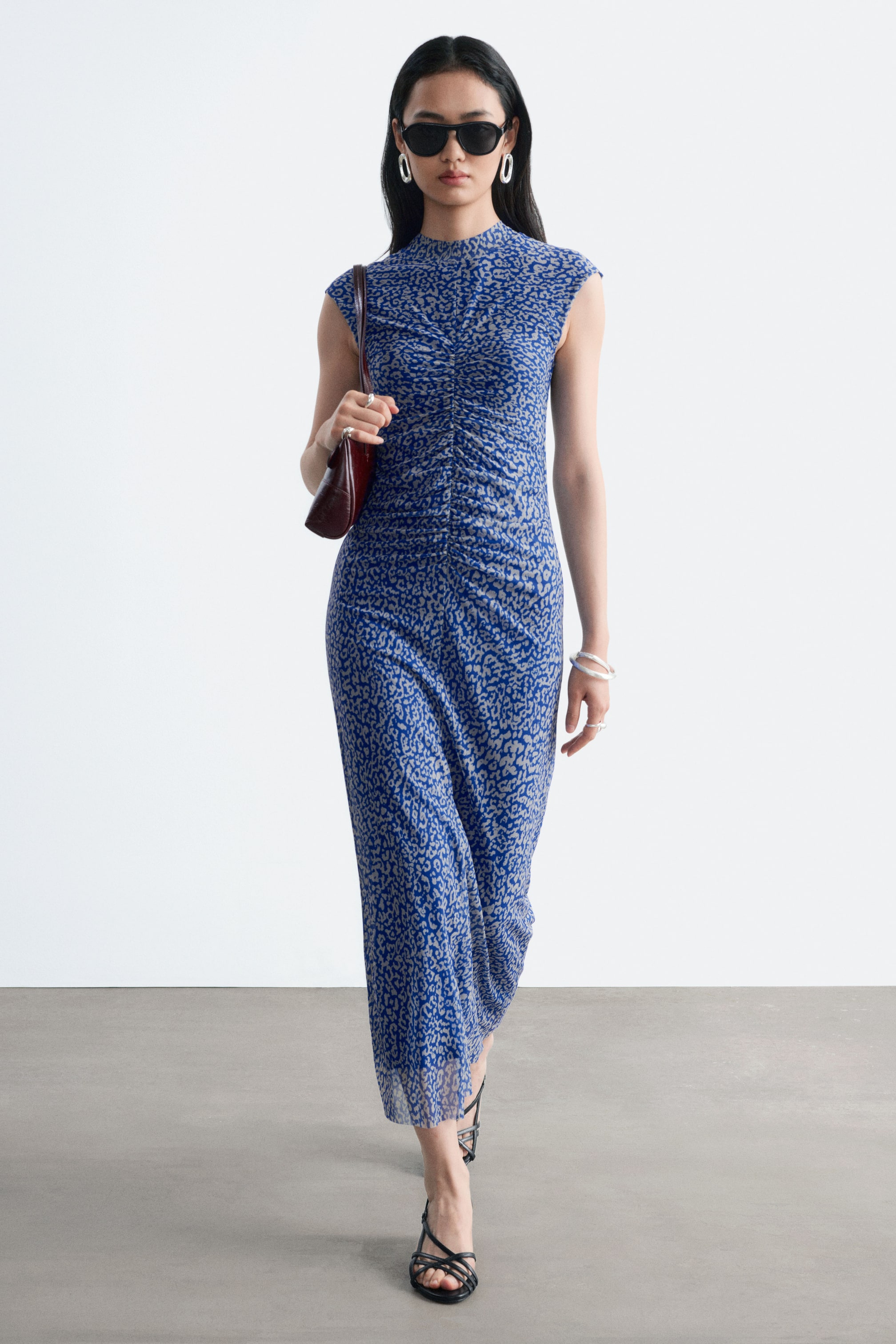 Größeres Bild ansehen: Ein blau-grau gemustertes Jerseykleid mit Stehkragen und vertikalen Raffungen in der vorderen Mitte wird von dem Model getragen. Das ärmellose Midikleid ist mit schwarzen Sandalen mit Riemchen und einer dunkelbraunen Umhängetasche kombiniert.