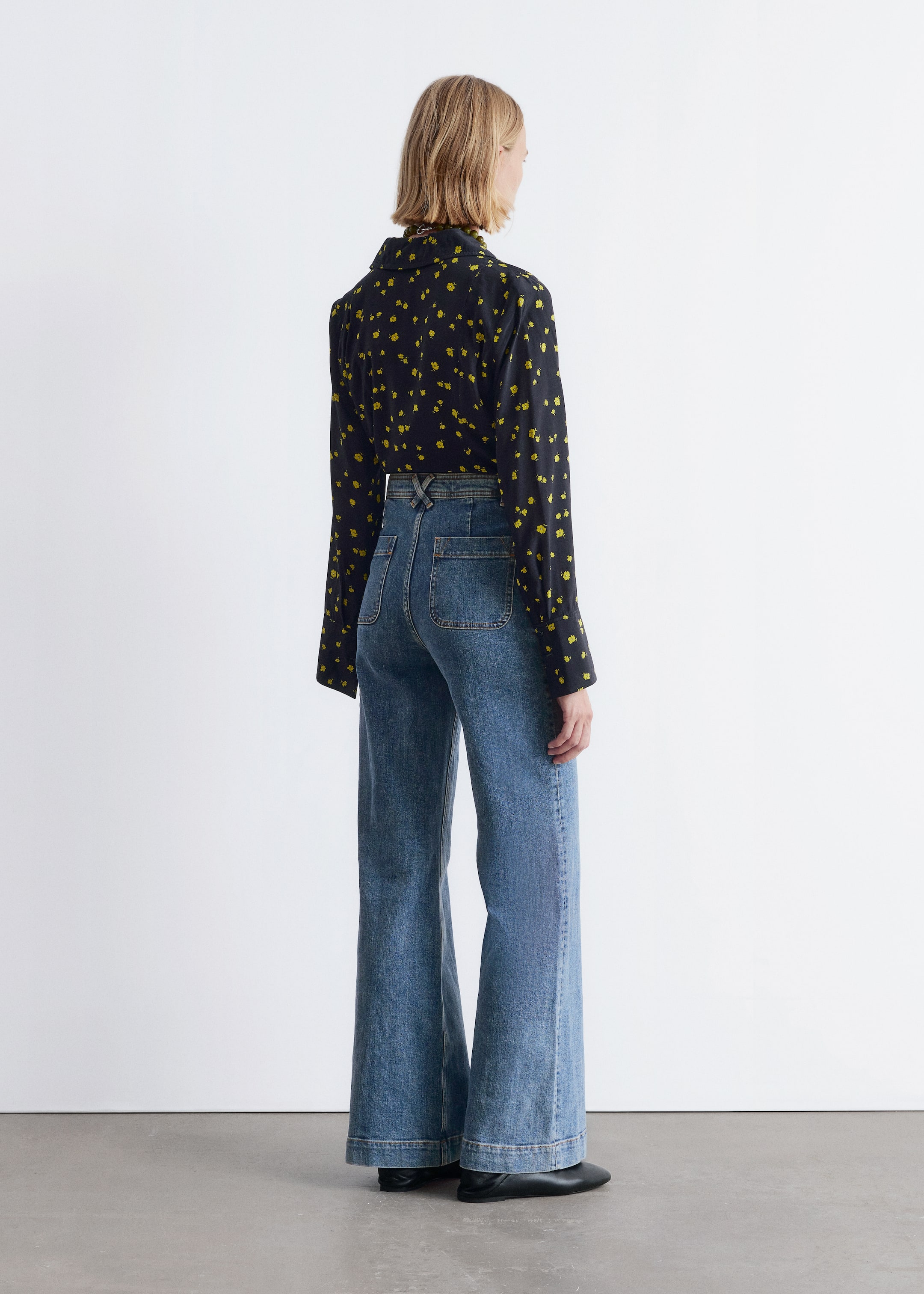 Größeres Bild anzeigen: Blaue Denim-High-Waist-Bootcut-Jeans werden vom Model getragen, von hinten betrachtet. Die Jeans verfügen über sichtbare aufgesetzte Taschen auf der Rückseite, abgestepptes Detail, Gürtelschlaufen und ein V-förmiges Detail am hinteren Bund, kombiniert mit einem dunklen Langarmhemd.
