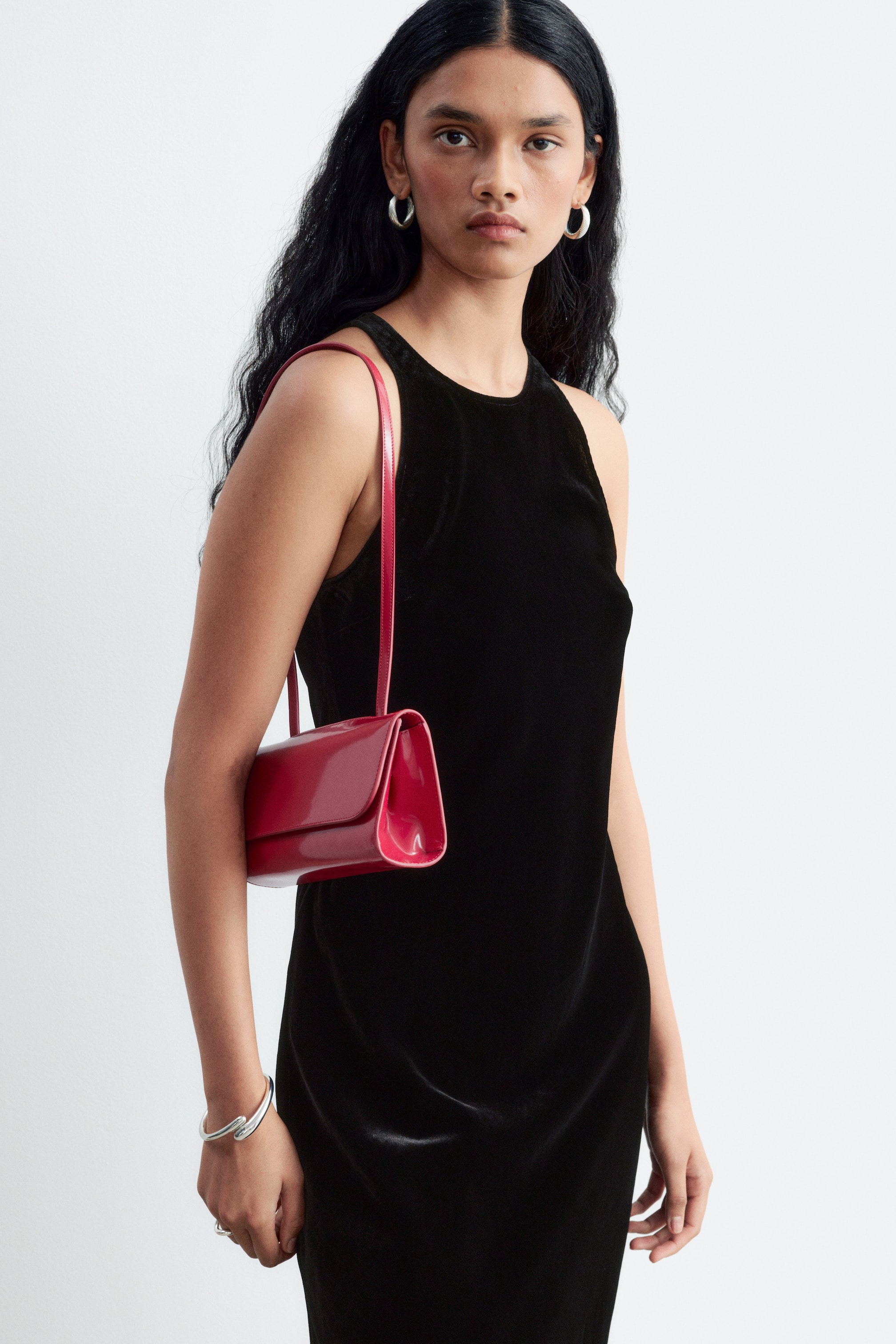 View larger image: A structured, glossy red shoulder bag is being worn on the shoulder by the model. The bag features a clean, rectangular silhouette with a front flap and a slim shoulder strap, styled against a black sleeveless dress.