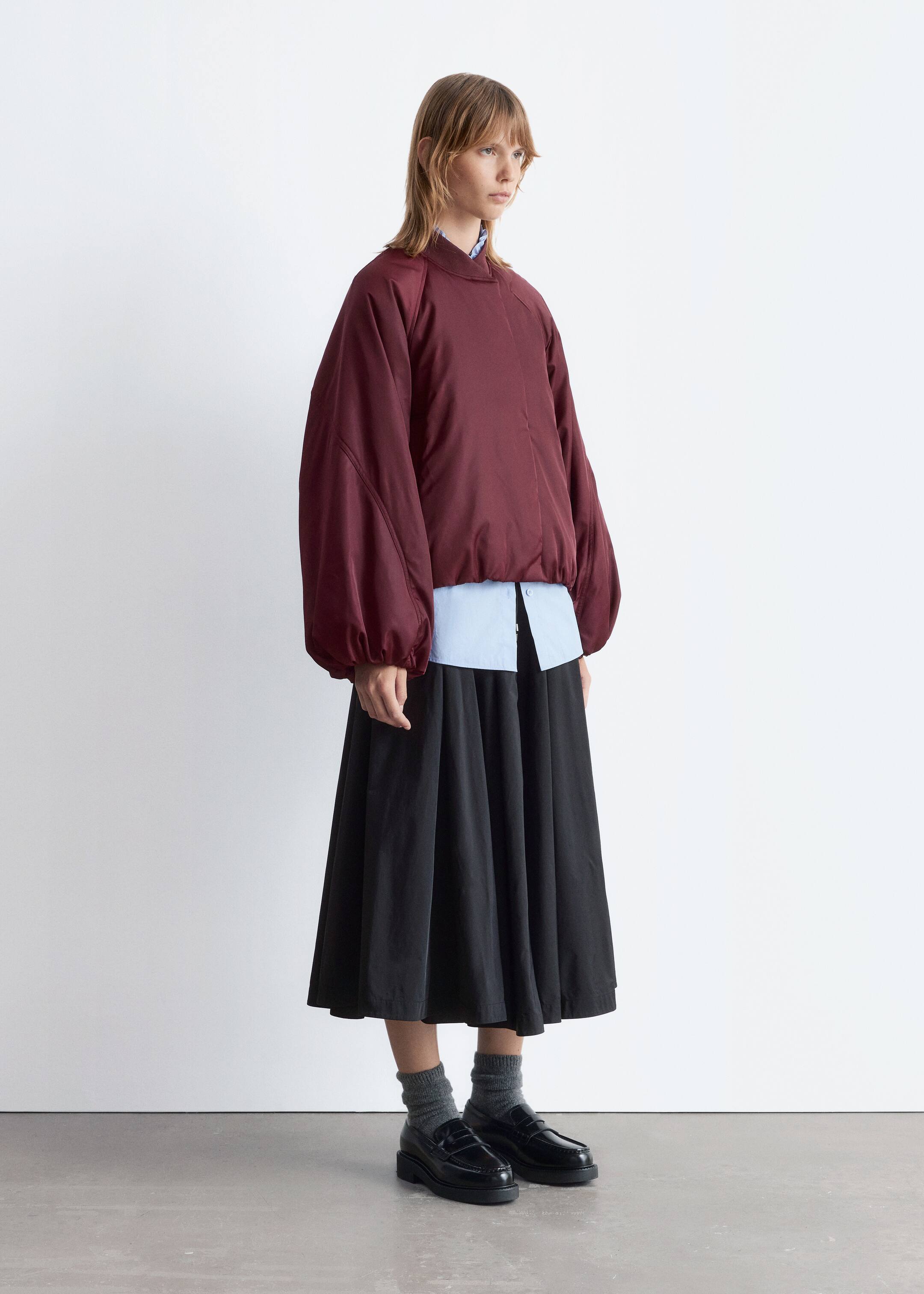 Visualizza immagine più grande: Un top color bordeaux in stile bomber, caratterizzato da maniche raglan voluminose raccolte ai polsini e un orlo elasticizzato, è indossato sopra una camicia azzurra con colletto. L'ensemble è presentato da una vista leggermente angolata, mostrando il top abbinato a una gonna nera a trapezio che cade a metà polpaccio, e completato con calzini grigi e mocassini neri massicci.