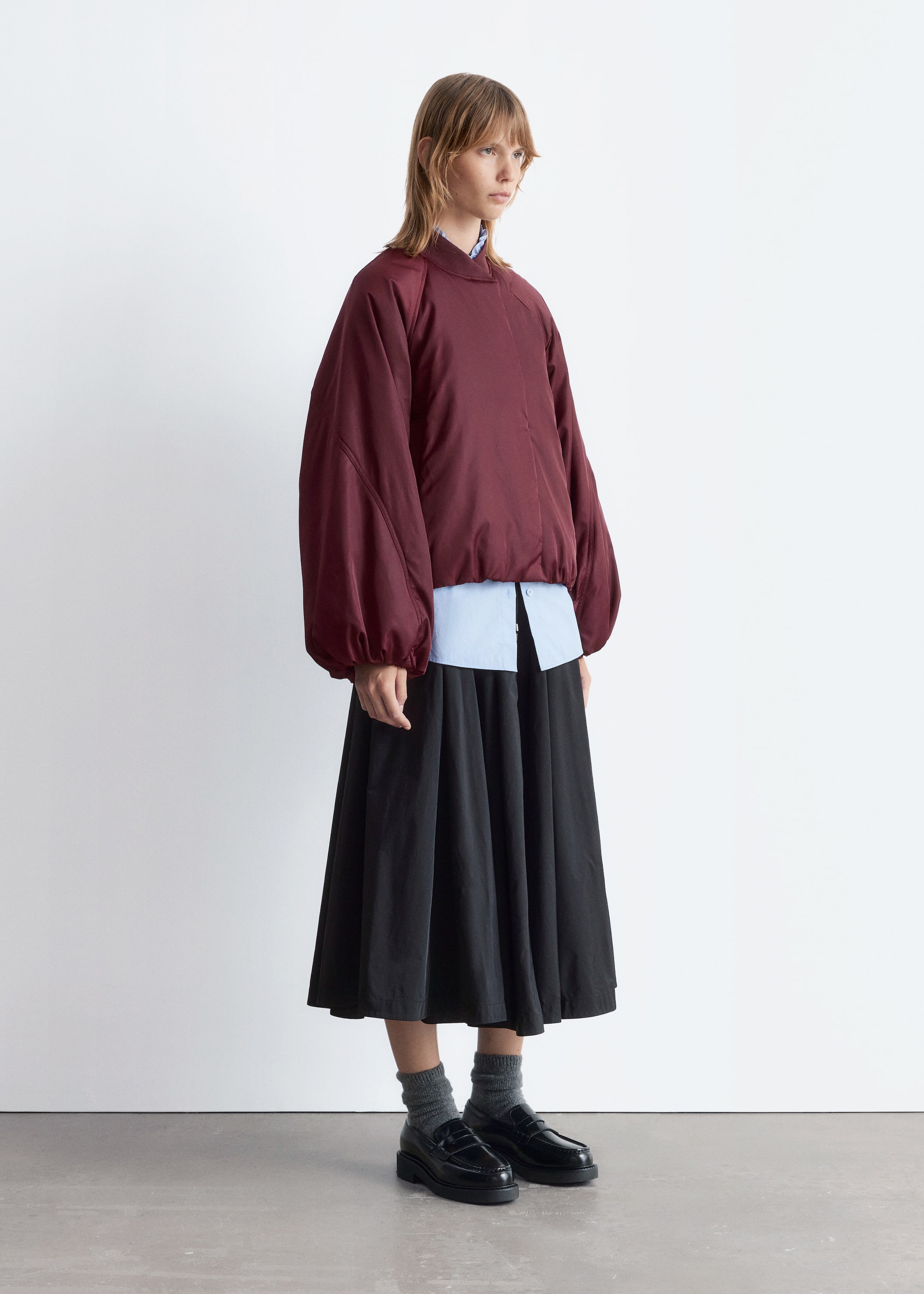 Visualizza immagine più grande: Un top color bordeaux in stile bomber, caratterizzato da maniche raglan voluminose raccolte ai polsini e un orlo elasticizzato, è indossato sopra una camicia azzurra con colletto. L'ensemble è presentato da una vista leggermente angolata, mostrando il top abbinato a una gonna nera a trapezio che cade a metà polpaccio, e completato con calzini grigi e mocassini neri massicci.