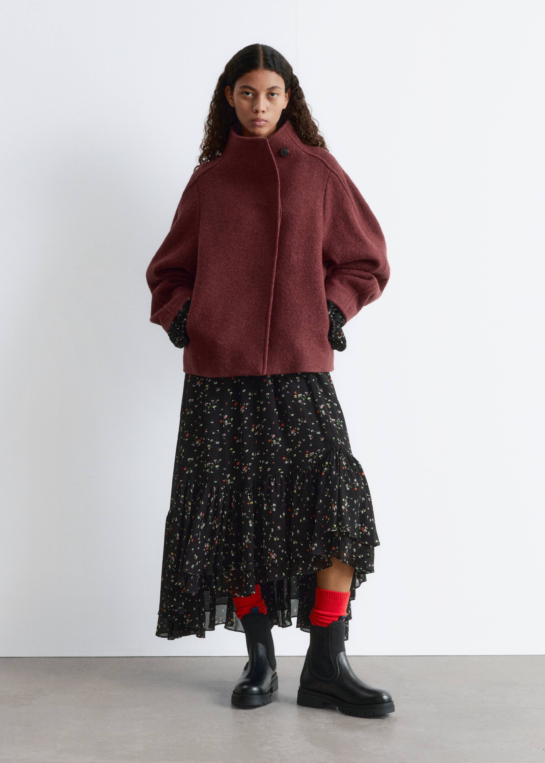 Grösseres Bild ansehen: Eine dunkelrote, gefilzte Jacke wird in der Vorderansicht präsentiert. Sie zeigt eine übergroße, verkürzte Silhouette mit einem hohen Stehkragen, der mit einem sichtbaren Knopf am Hals befestigt wird, und einem verdeckten Frontverschluss, während voluminöse lange Ärmel, die bis zu den Handgelenken reichen, mit schwarz gemusterten Ärmeln, die darunter hervorlugen.