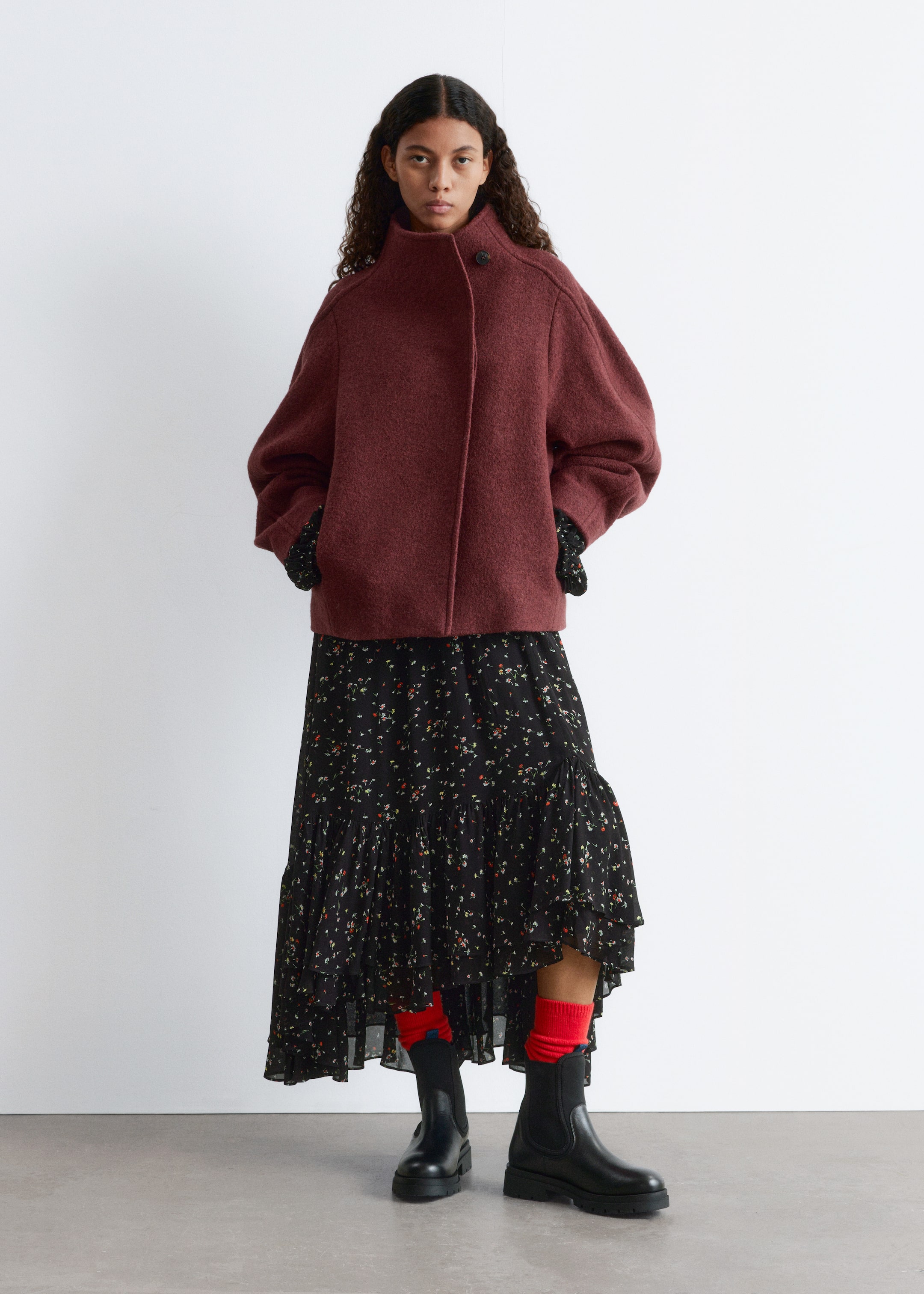 Ingrandisci l'immagine: Una giacca rosso scuro in feltro viene presentata da una vista frontale. Mostra una silhouette oversize e cortata, con un colletto alto che si allaccia con un bottone visibile al collo e una chiusura frontale nascosta, mentre le maniche lunghe voluminose si estendono fino ai polsi con le maniche con motivo nero che sbucano da sotto.