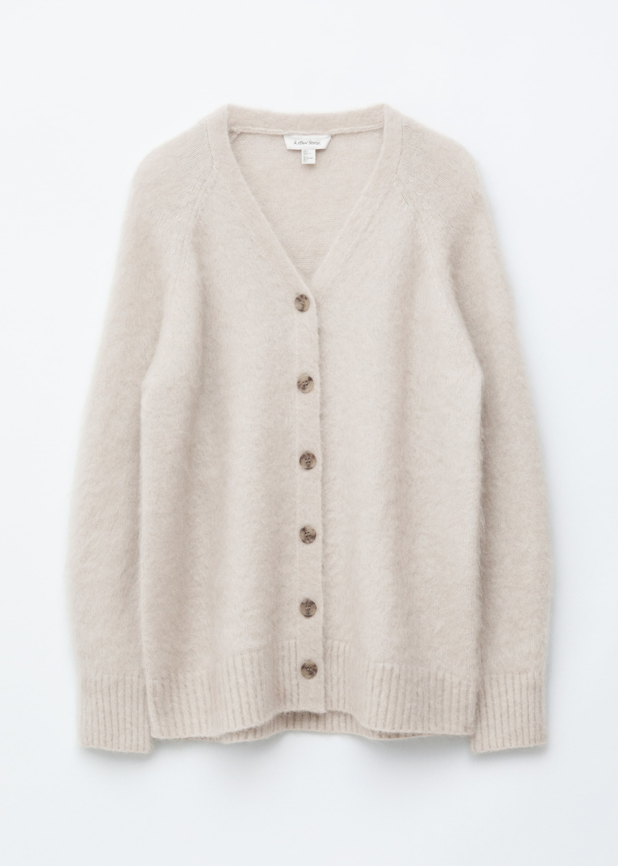 Visualizza immagine più grande: Un cardigan beige chiaro mostra la parte anteriore, caratterizzato da una texture a maglia pelosa, uno scollo a V, maniche raglan e sei bottoni rotondi marroni lungo il centro, rifinito con polsini e orlo a coste.