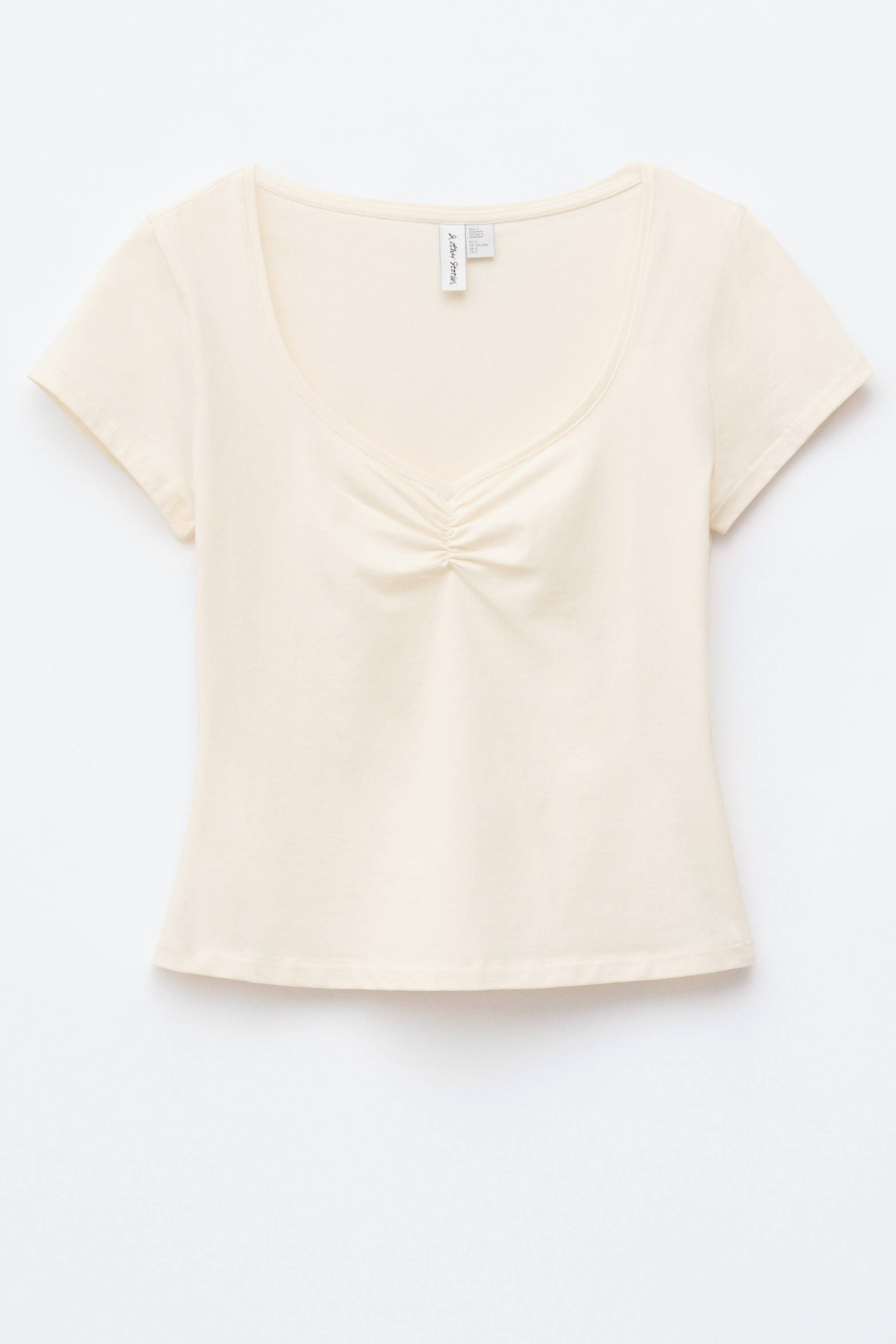 Se større billede: En cremefarvet, slim-fit top vises forfra, med en rund halsudskæring, korte ærmer og en rynkedetalje i midten af brystet.
