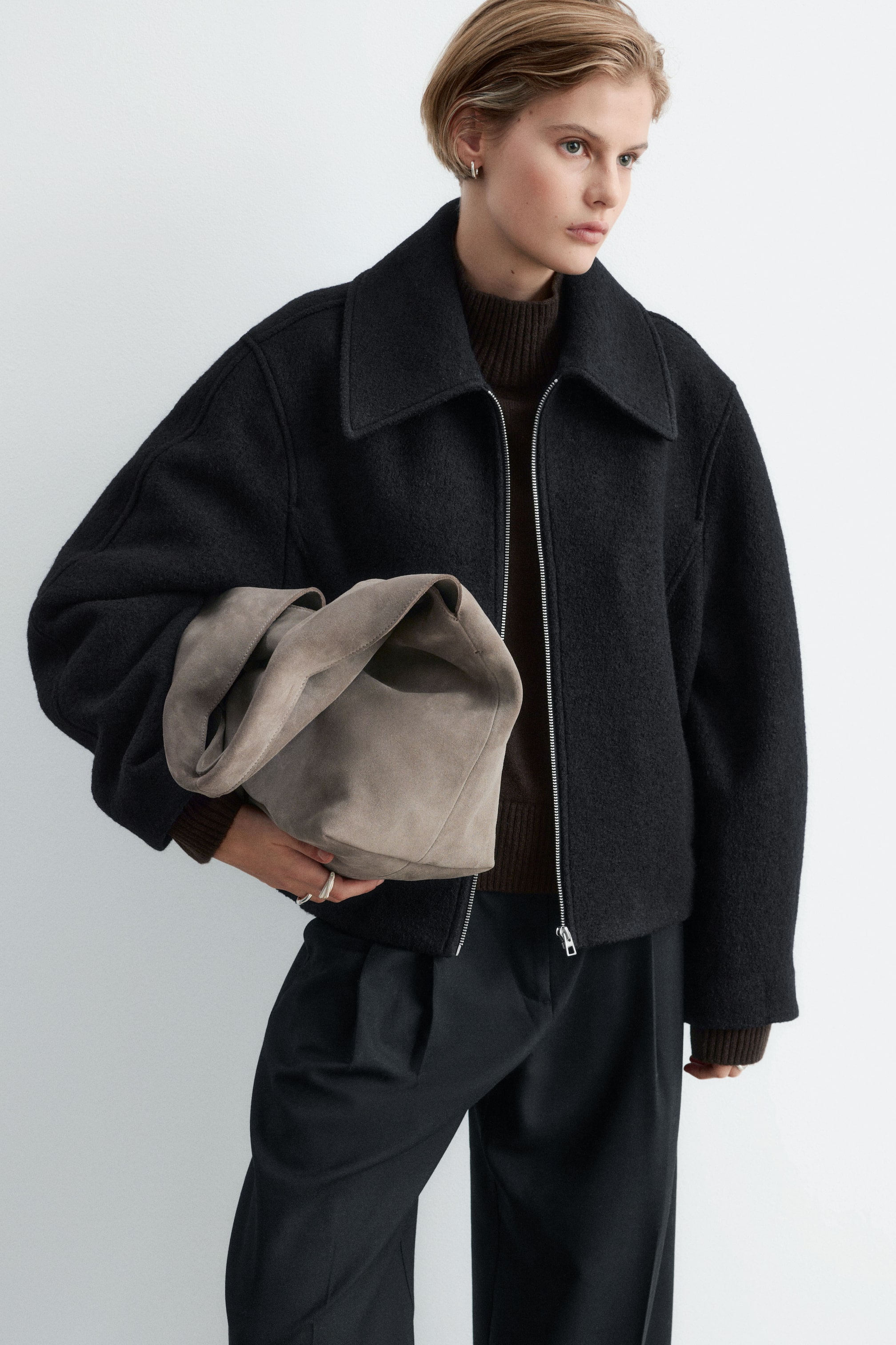 Agrandir l'image: Un sac fourre-tout en cuir suédé fendu gris à la silhouette souple est porté sous le bras par le mannequin. Le sac est maintenu contre une veste noire zippée, mettant en valeur sa texture douce et sa forme décontractée.