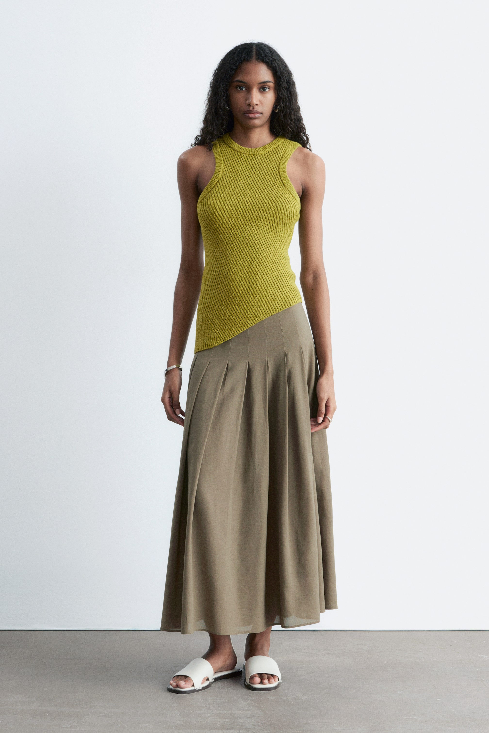 View larger image: The model is wearing a khaki green midi skirt, characterised by a flared A-line silhouette and fixed pleats. It is paired with a lime green ribbed vest top and white flat sandals.