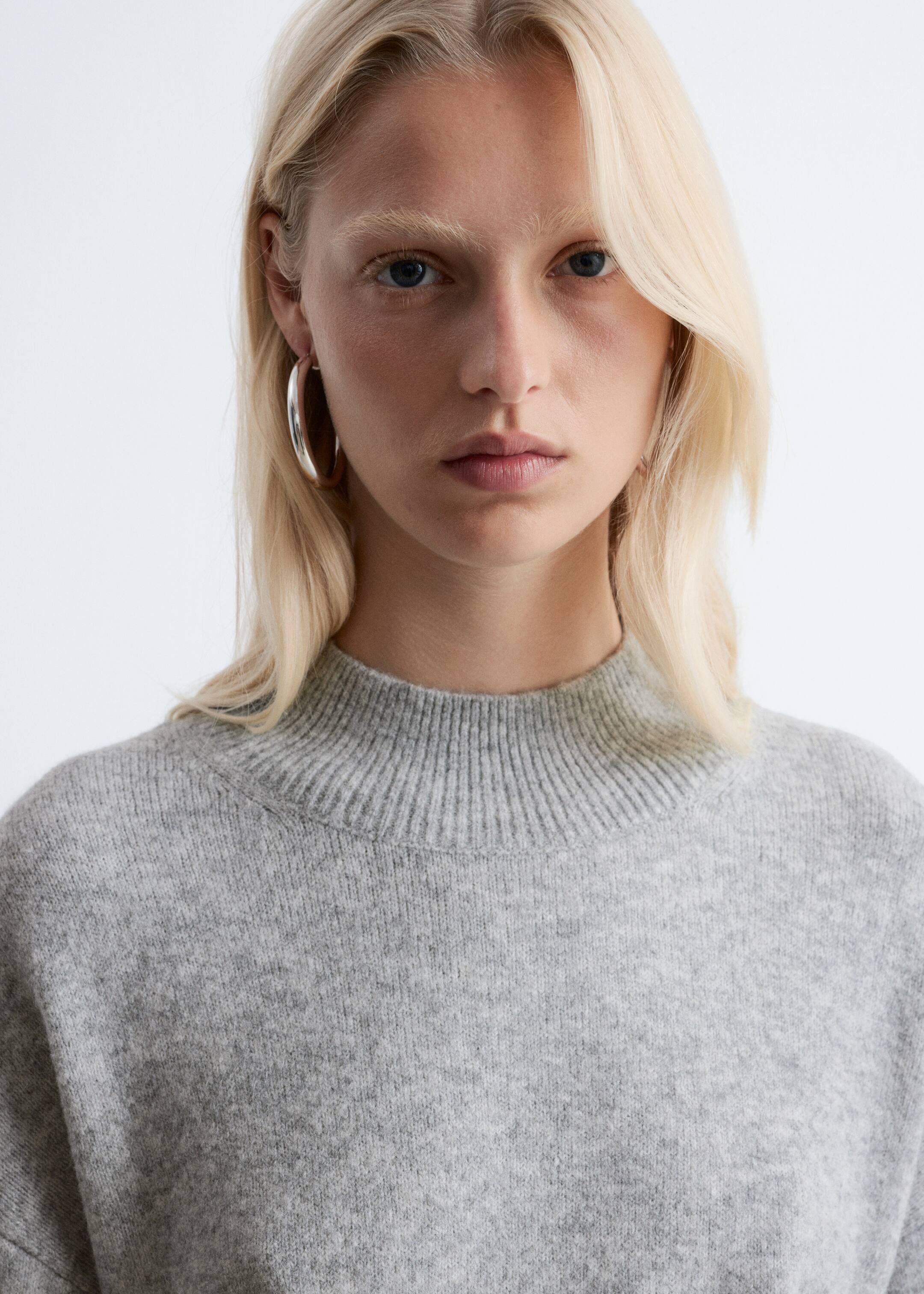 Größeres Bild anzeigen: Ein hellgrauer Strickpullover wird in einer frontalen Nahaufnahme gezeigt. Er verfügt über einen gerippten Stehkragen und eine weiche, leicht flauschige Textur über der sichtbaren oberen Brust und den Schultern.