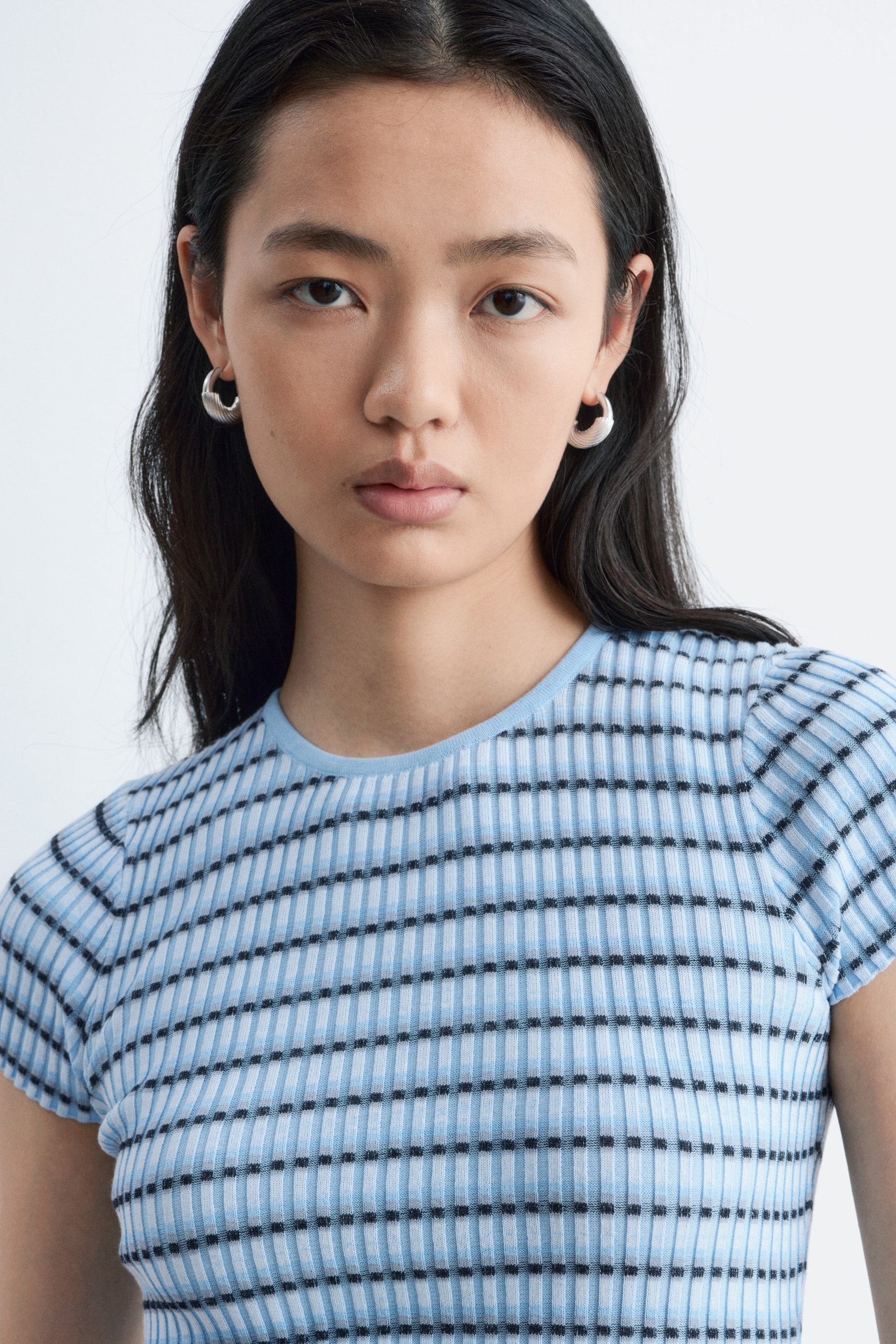 Visa större bild: En ljusblå och vit ribbstickad topp med rund hals och korta ärmar visas från en framåtvänd vinkel, med ett randigt mönster av horisontella ljusblå, vita och små svarta fyrkantiga band.