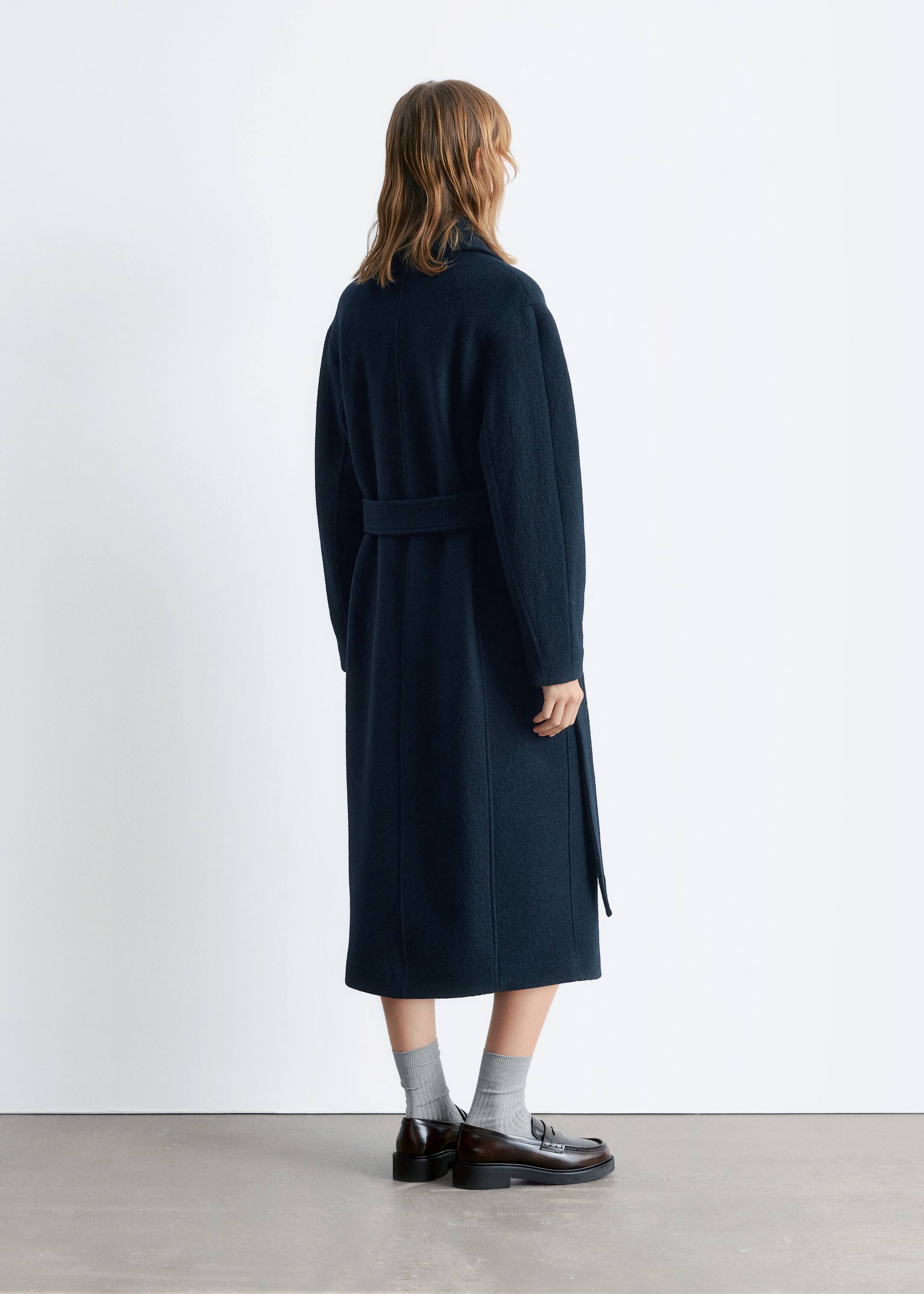 Agrandir l'image: Un long manteau bleu foncé est présenté de dos et de profil légèrement latéral. Il présente une coupe décontractée avec des manches longues et une ceinture assortie nouée à l'arrière, cintrant la taille.