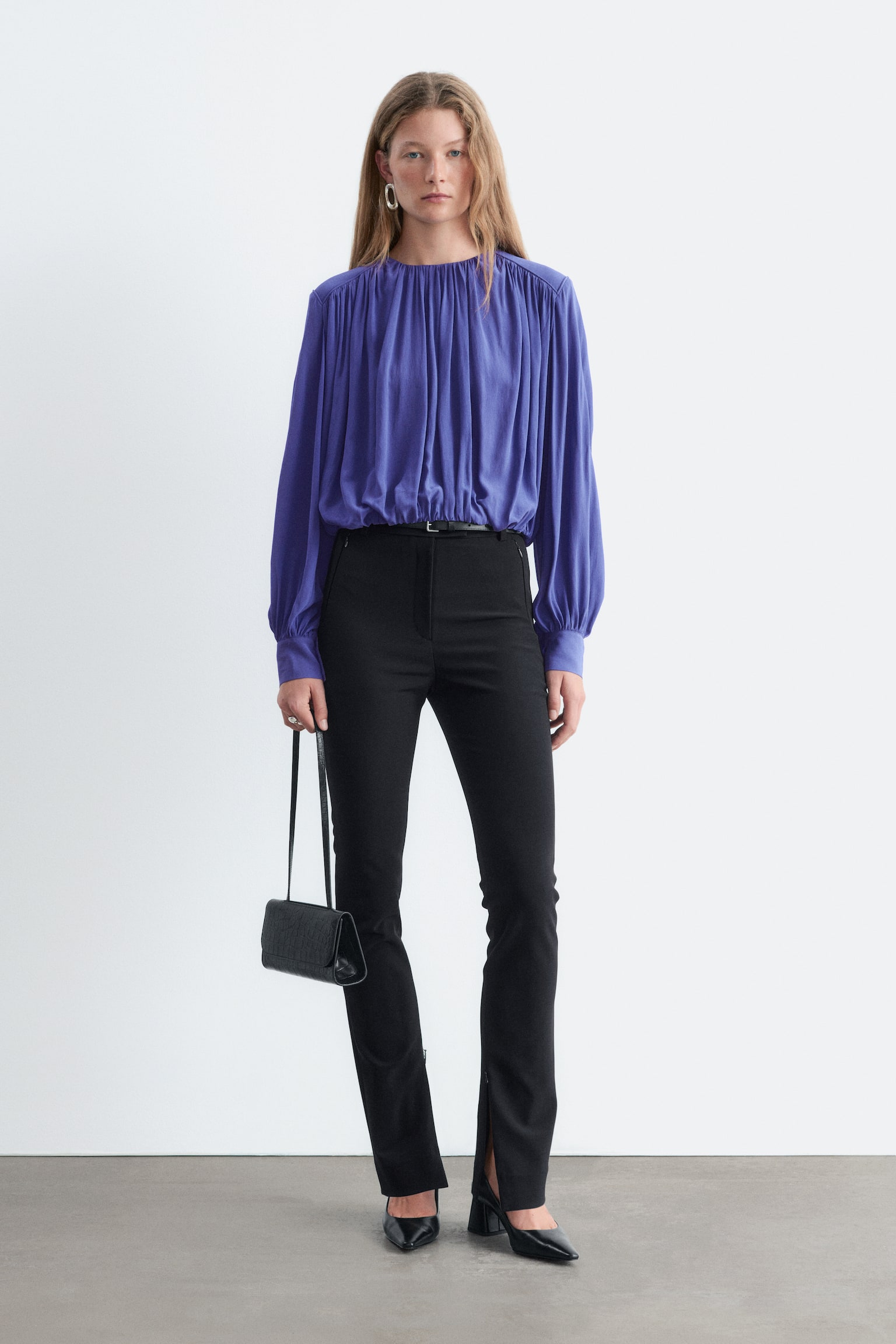 Pantalonii negri slim-fit sunt prezentaÈi din faÈÄ, avÃ¢nd buzunare laterale cu fermoar, gÄici pentru curea Èi fermoare pe cusÄtura interioarÄ la tivul de jos, care creeazÄ deschideri subtile la nivelul cracilor. Pantalonii sunt stilizaÈi cu o bluzÄ mov cu mÃ¢necÄ lungÄ, bÄgatÄ Ã®n pantaloni, Èi pantofi cu toc negri pentru a evidenÈia designul cu tiv despicat. - 1