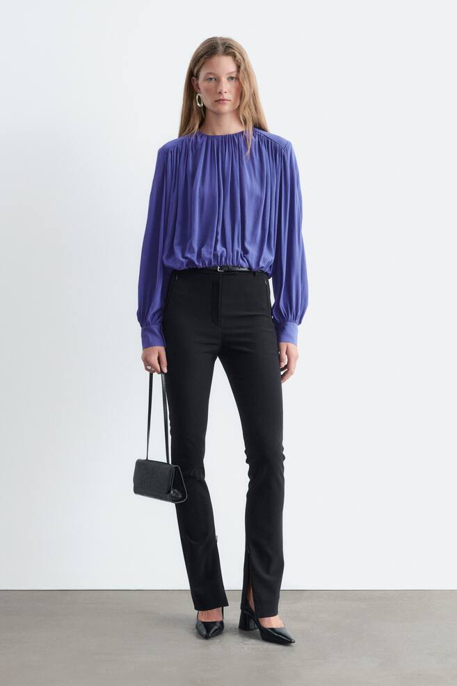 Slim-fit black trousers are shown from the front, featuring zipped side pockets, belt loops, and inside seam zips at the bottom hem that create subtle leg openings. The trousers are styled with a tucked-in purple long-sleeved blouse and black heels to highlight the split-hem design. - 1