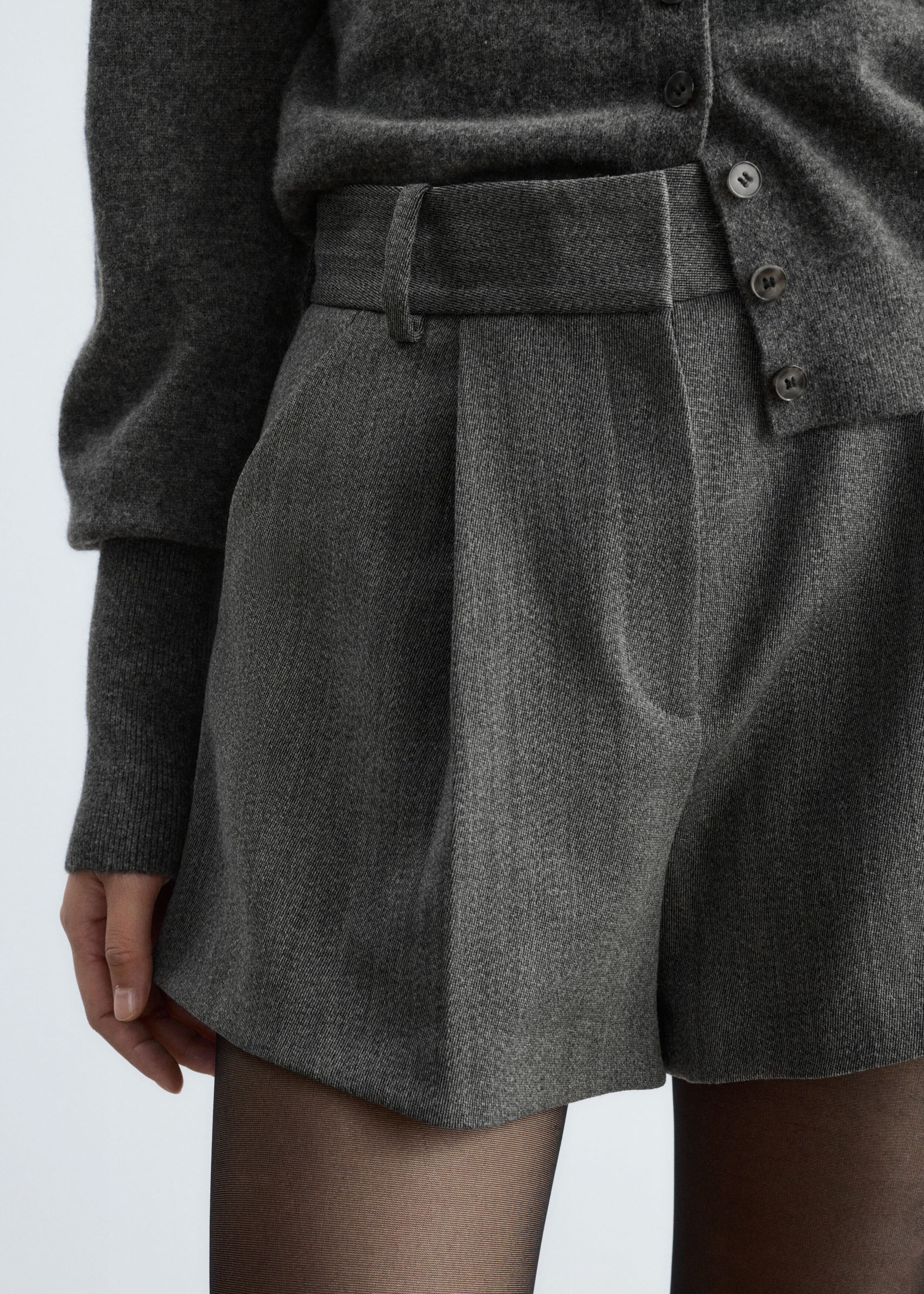 These high-waisted gray wool-blend shorts with front pleats and belt loops are worn by the model. The shorts are styled with a matching gray button-up cardigan and sheer black tights.
