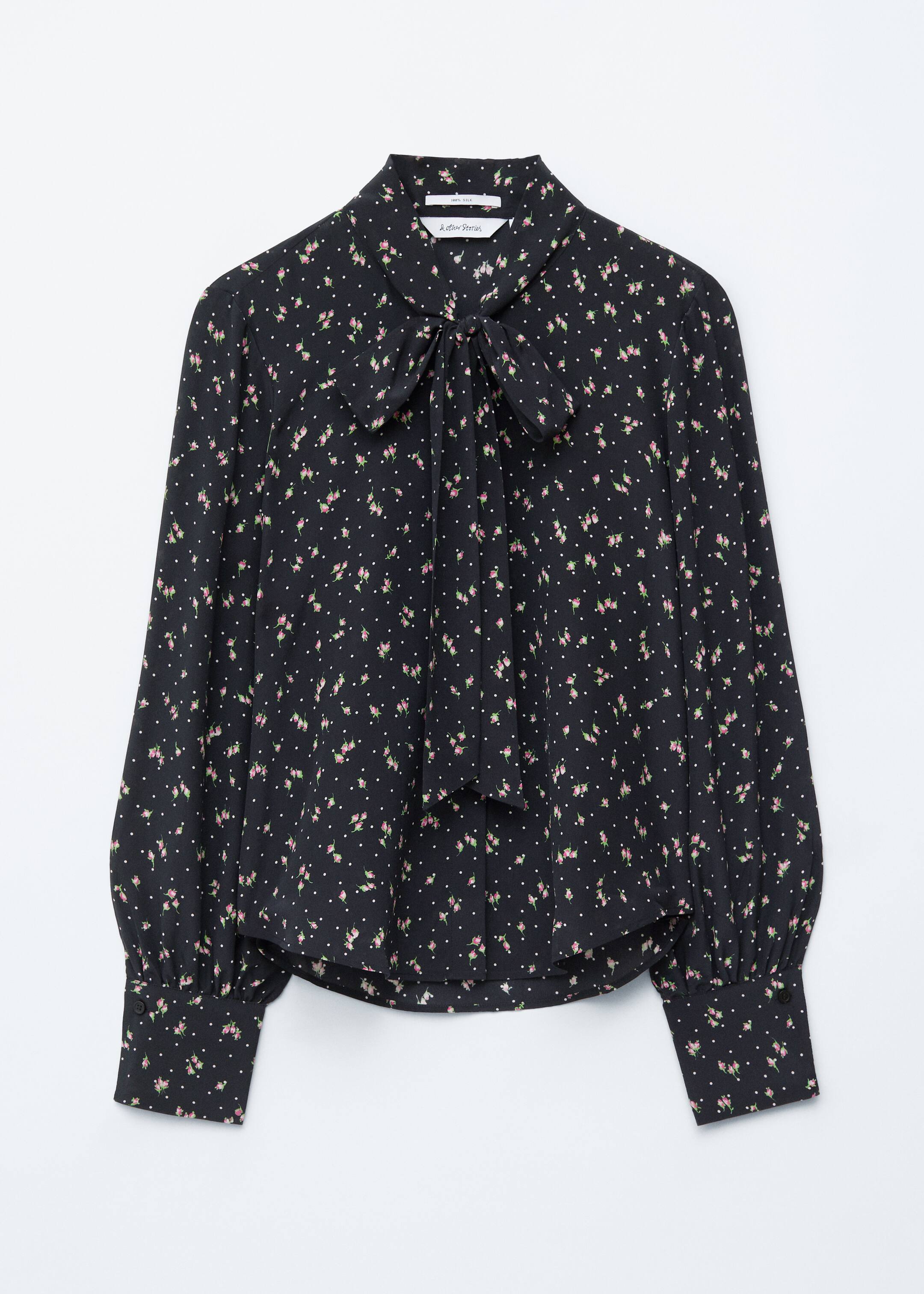 Größeres Bild anzeigen: Eine schwarze Bluse mit rosa Blumen und weißen Punkten wird von vorne präsentiert, mit einem Kragen, der zu einer Schleife gebunden ist, einer verdeckten Knopfleiste und langen, voluminösen Ärmeln, die mit breiten, geknöpften Manschetten enden.