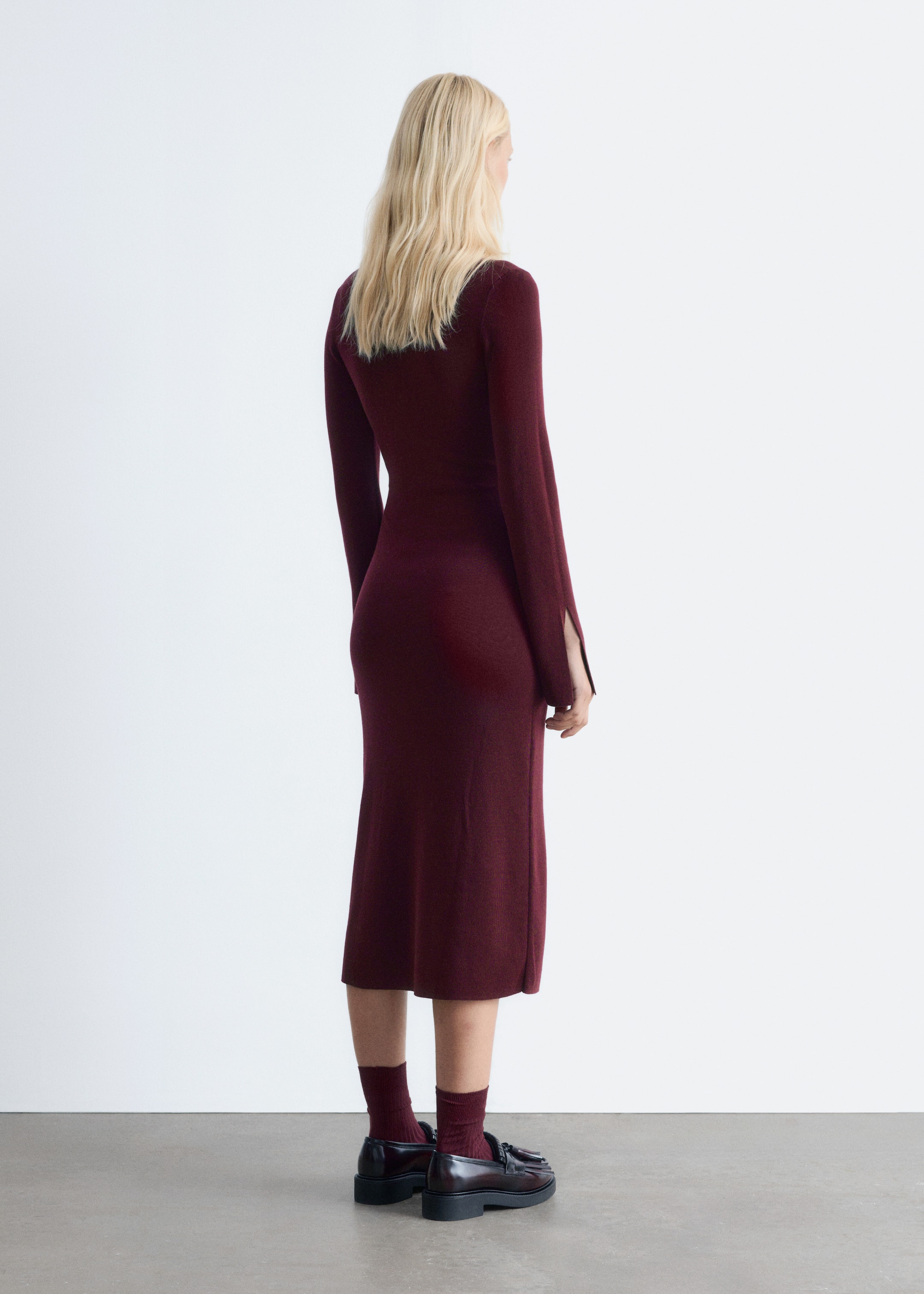 Ingrandisci l'immagine: Un abito midi a maglia di colore rosso scuro con una vestibilità aderente e maniche lunghe è indossato dalla modella. L'abito presenta spacchi sui polsini delle maniche e la modella indossa anche calzini coordinati e mocassini scuri.