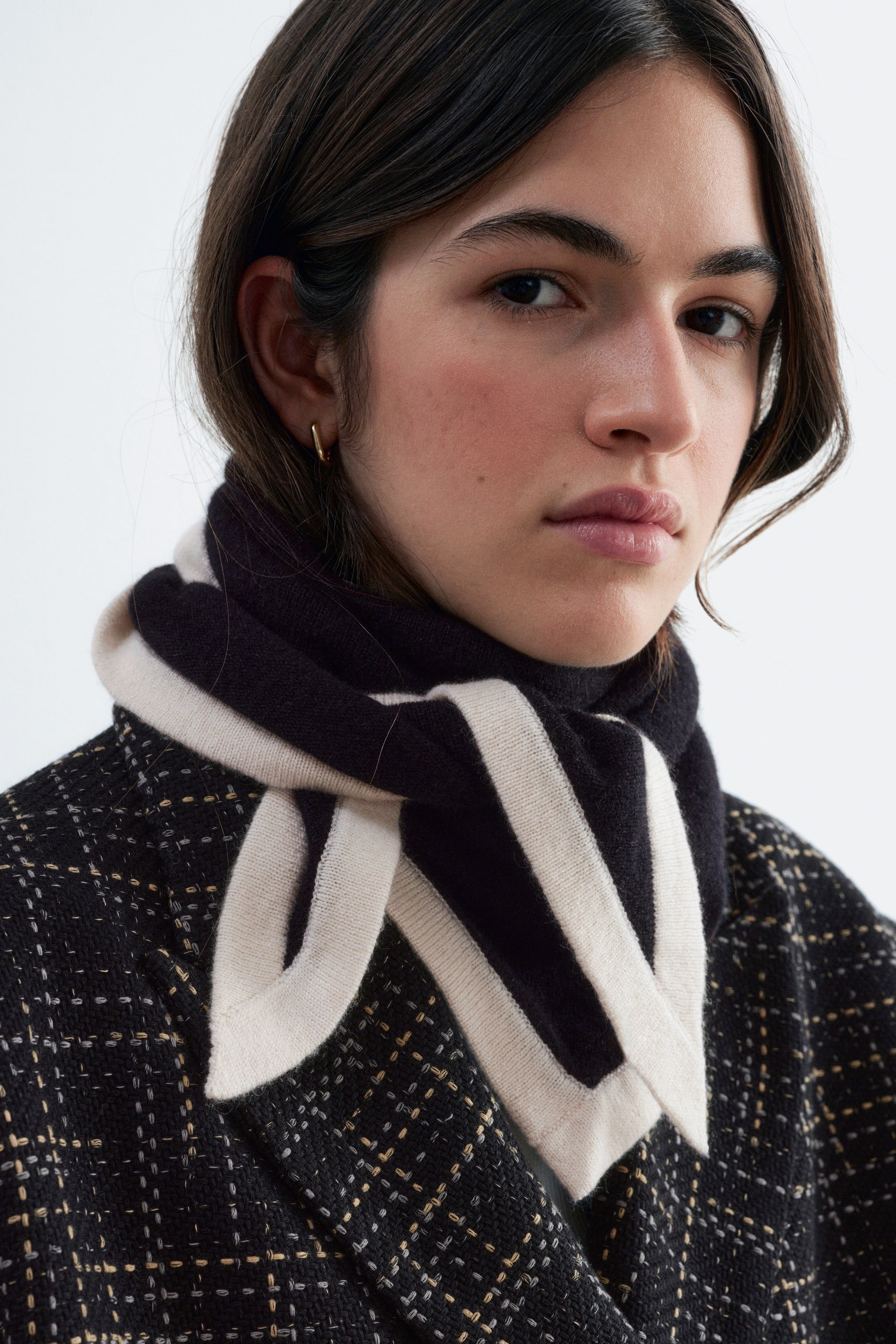 View larger image: A silk-cashmere triangular scarf in black with a contrasting cream border is being worn by the model, wrapped closely around the neck and tied in a loose knot at the front. The scarf is layered over a dark, textured tweed jacket.