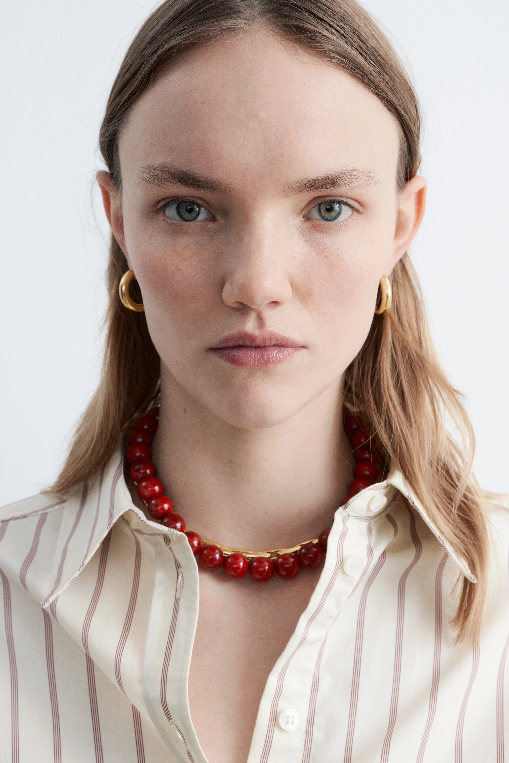 Grösseres Bild ansehen: Ein kurzes rotes Steinperlen-Halsband mit einem goldfarbenen O-Ring-Verschluss wird vom Model getragen. Es liegt auf dem offenen Kragen eines hellen gestreiften Hemdes.