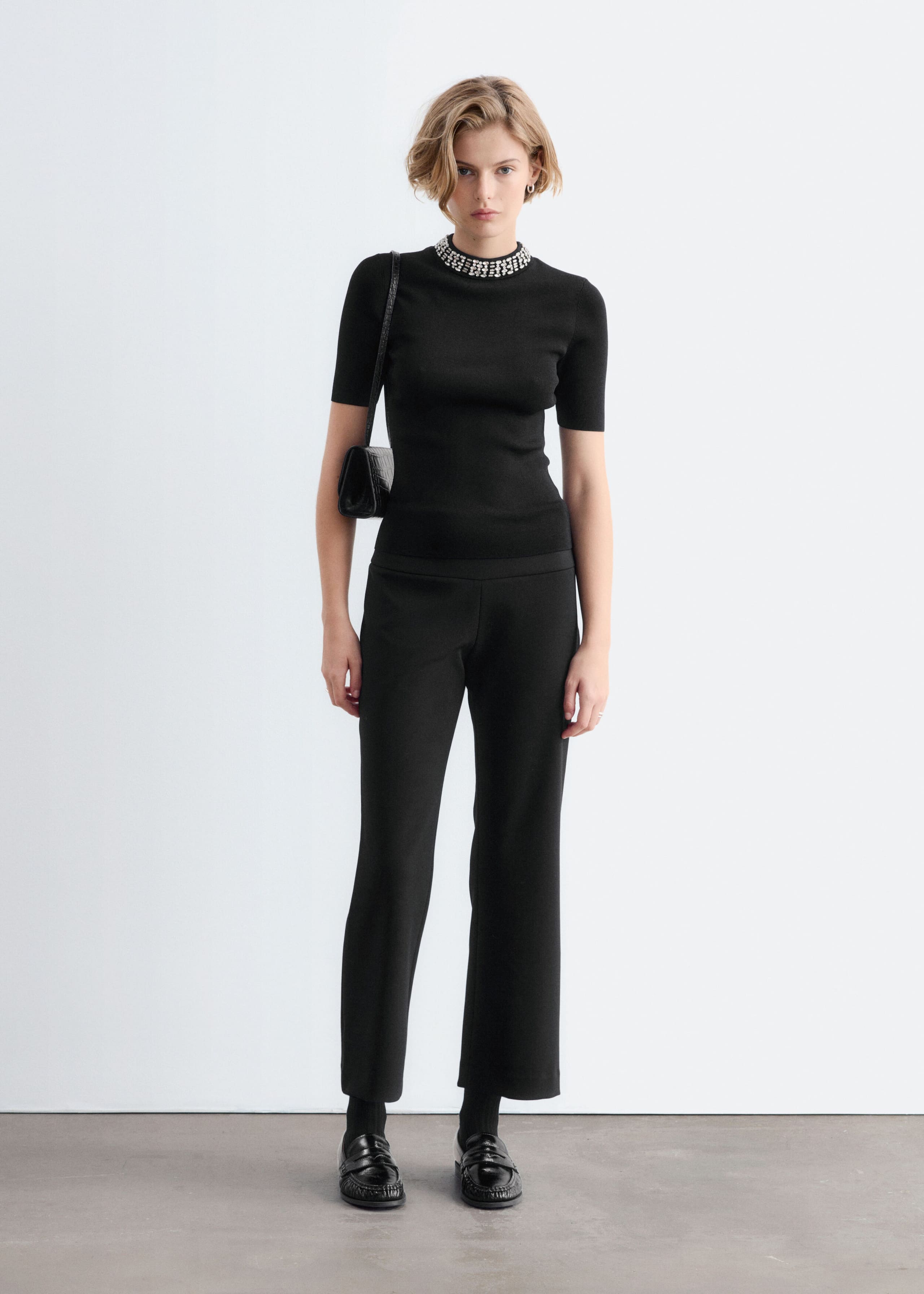 A black, short-sleeved fitted top featuring a mock neck embellished with rows of silver rhinestone beads is worn by the model. The top is styled with matching black trousers and a small black shoulder bag.