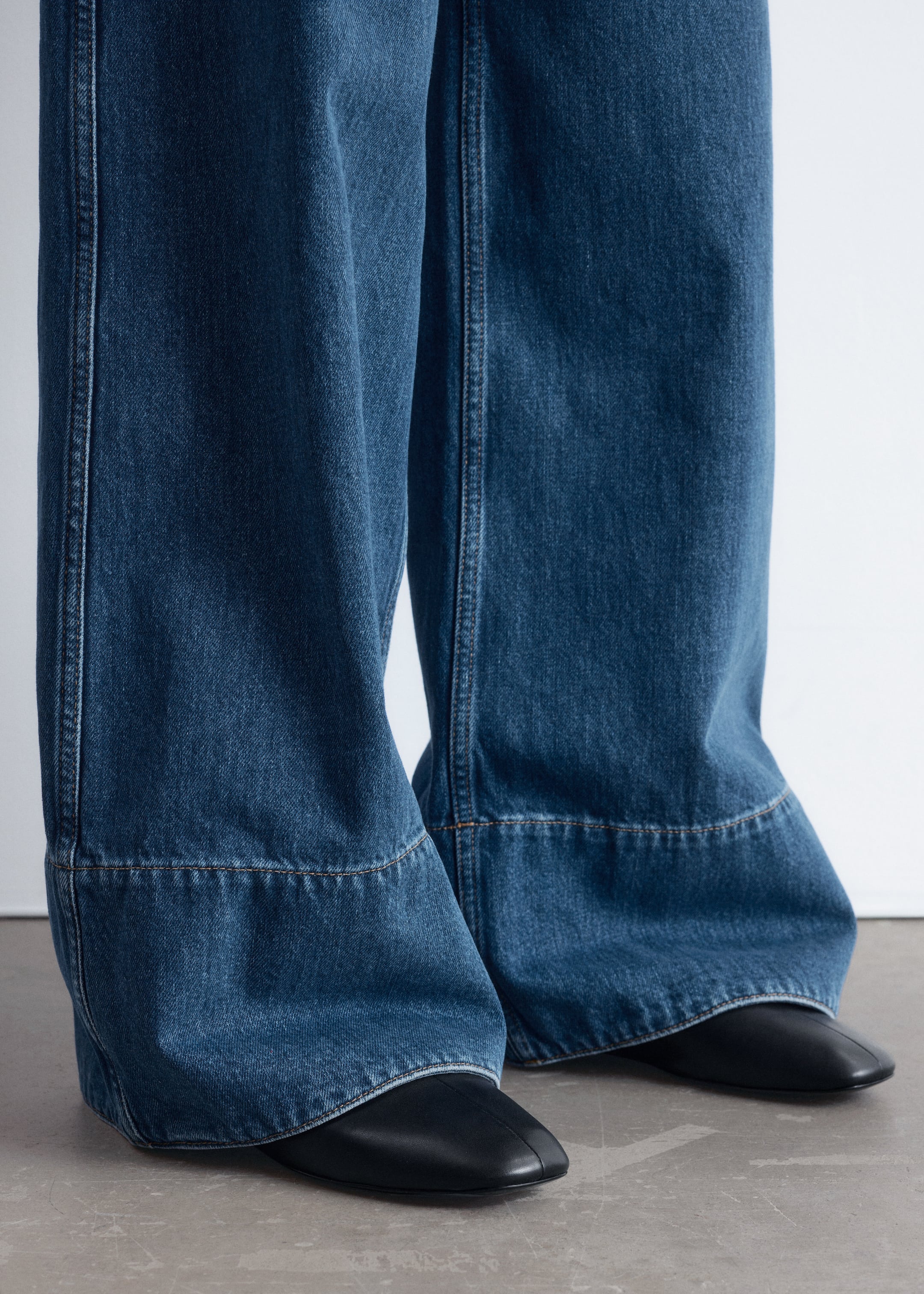 Grösseres Bild ansehen: Dunkelblaue Jeans mit weitem Bein werden getragen, die sichtbare, oben gestickte vertikale Nähte aufweisen. Eine markante, horizontal oben gestickte Naht bildet ein breites Panel am Saum, wobei die Jeans über schwarzen flachen Schuhen zusammenfällt.