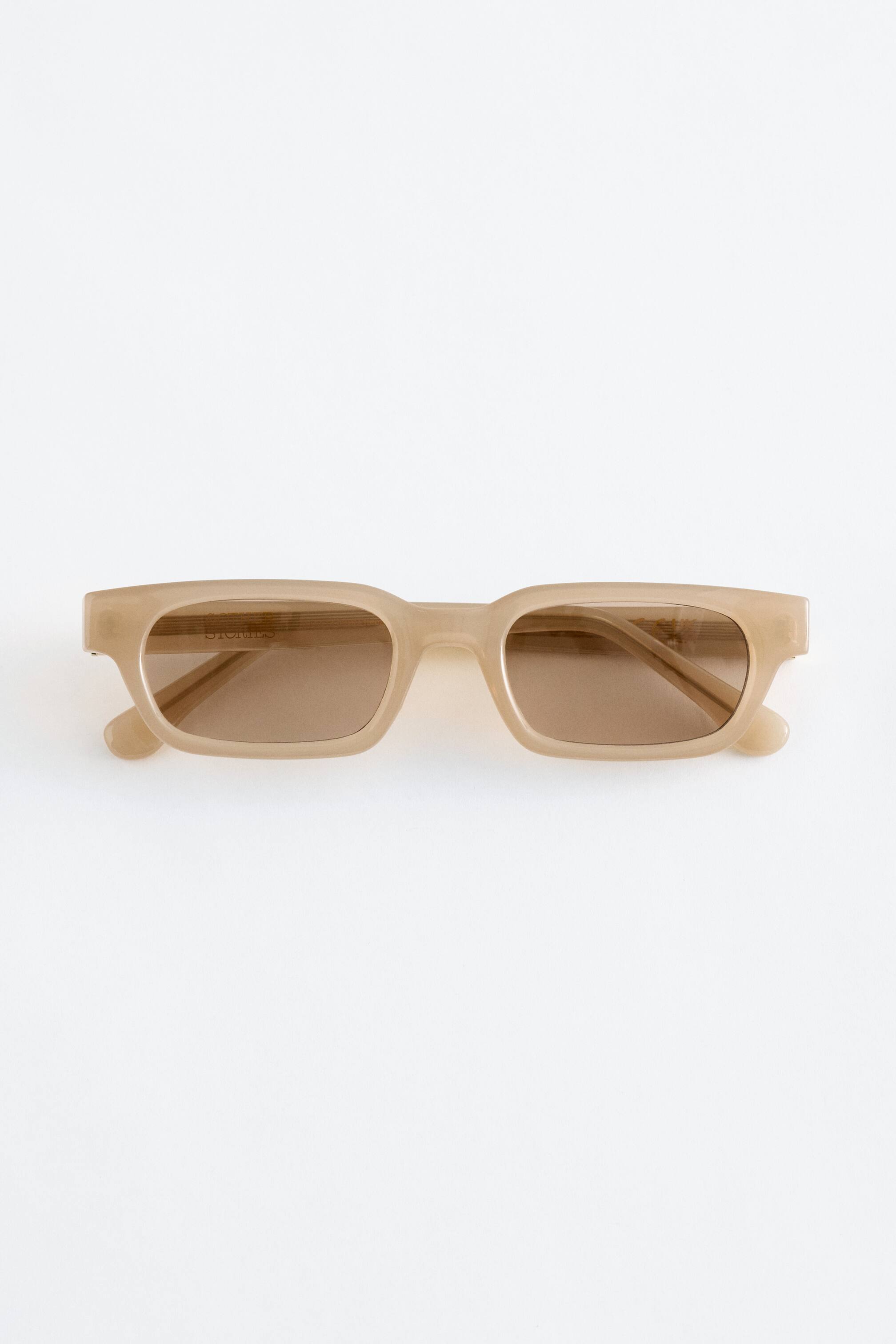 Grotere afbeelding bekijken: Smal omrande zonnebrillen met doorschijnende beige monturen en rechthoekige, bruin getinte glazen, die een vooraanzicht van het ontwerp presenteren.