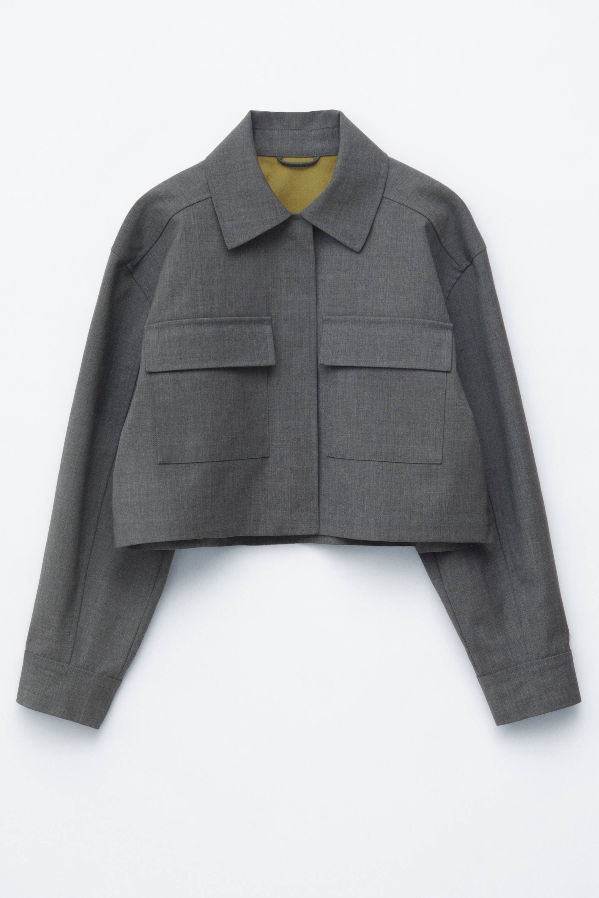 Visa större bild: En grå kort boxig jacka visas i sin FRAMSIDES vy, med en klassisk krage, dubbla bröstfickor med lock och synliga toppsömmar. Ett kontrasterande olivgrönt foder syns vid halsöppningen.