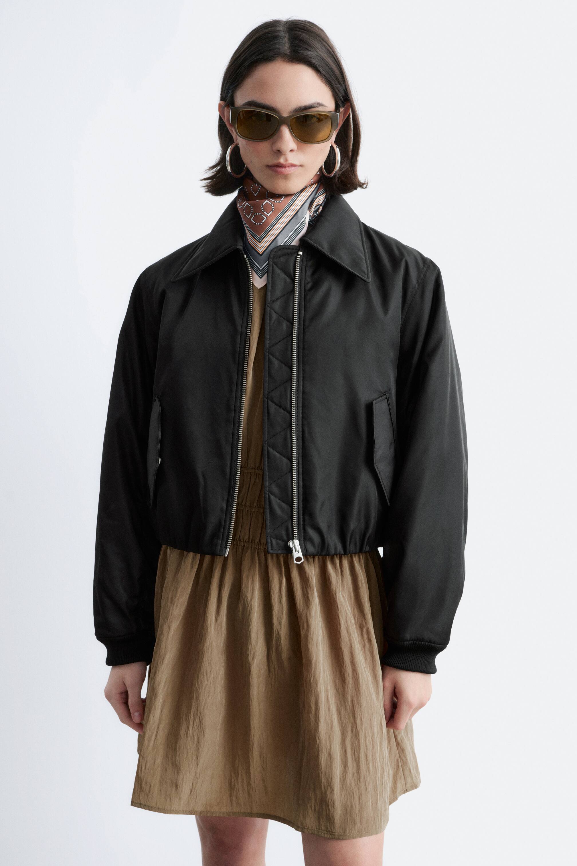 Преглед на по-голямо изображение: Черен скъсен бомбер яке, с остро яка, частично отворено закопчаване с цип, разкриващо диамантено капитонирана подплата, и джобове с кант, носено от модела върху светлокафява рокля. Под яката на якето се носи шарен шал.