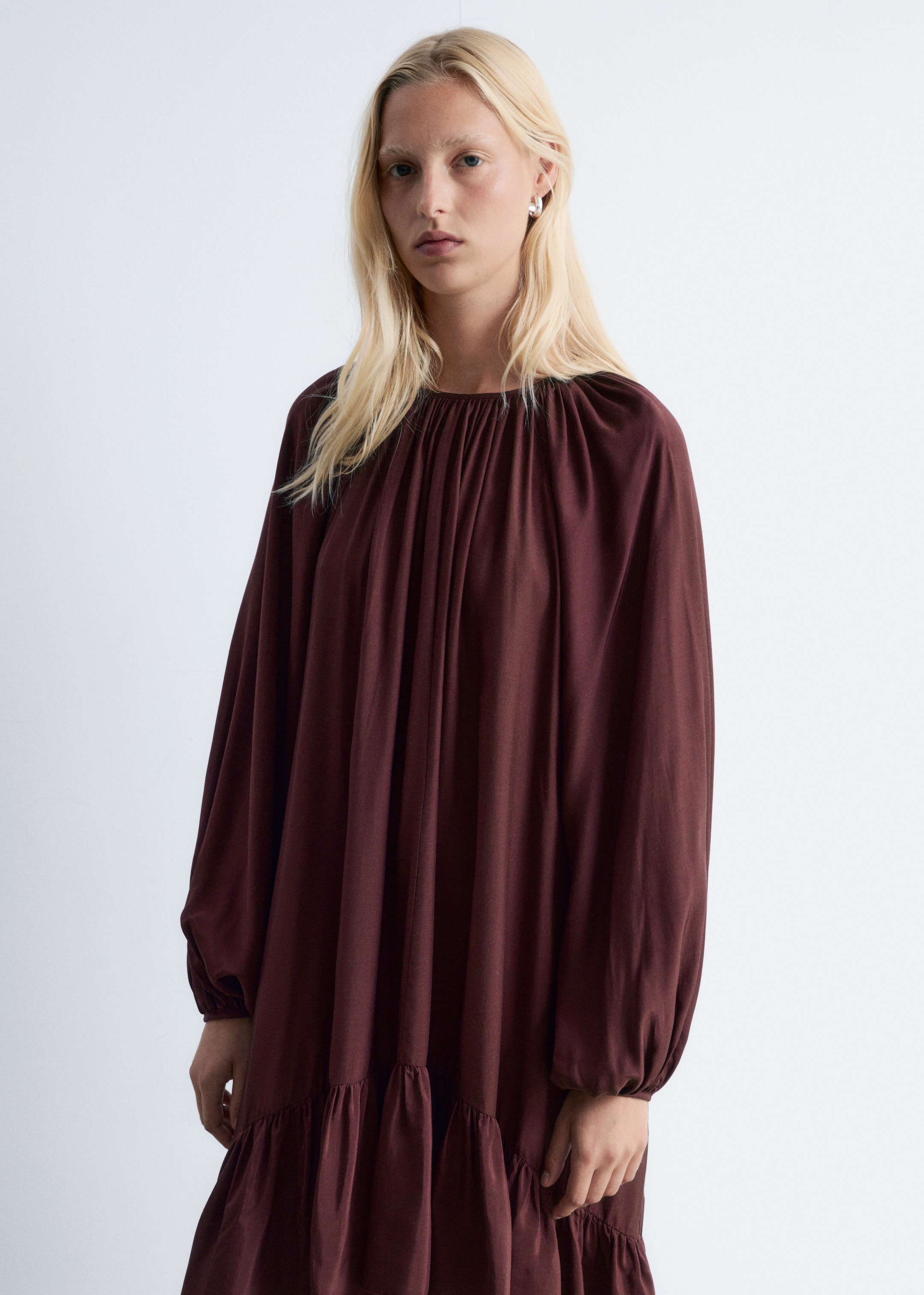Grösseres Bild ansehen: Ein dunkelbraunes Kleid wird vorwärts gerichtet vorgeführt. Es verfügt über einen hoch gerafften runden Ausschnitt, voluminöse lange Bischofsärmel mit gerafften Manschetten und eine lockere, fließende Silhouette mit einem gestuften, gerüschten Saum.
