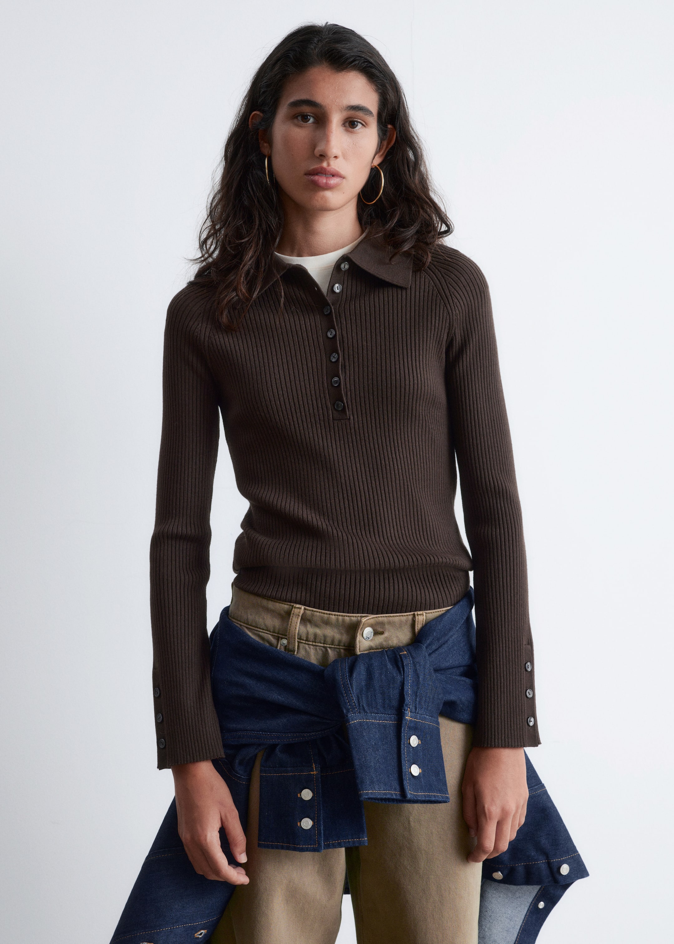 Grotere afbeelding bekijken: Een donkerbruine, slim-fit, geribbelde gebreide poloshirt met een klassieke kraag en een half knoopsluiting wordt gedragen door het model. Het shirt heeft lange mouwen met knopen aan de manchetten, en een wit ondershirt is zichtbaar onder de kraag.