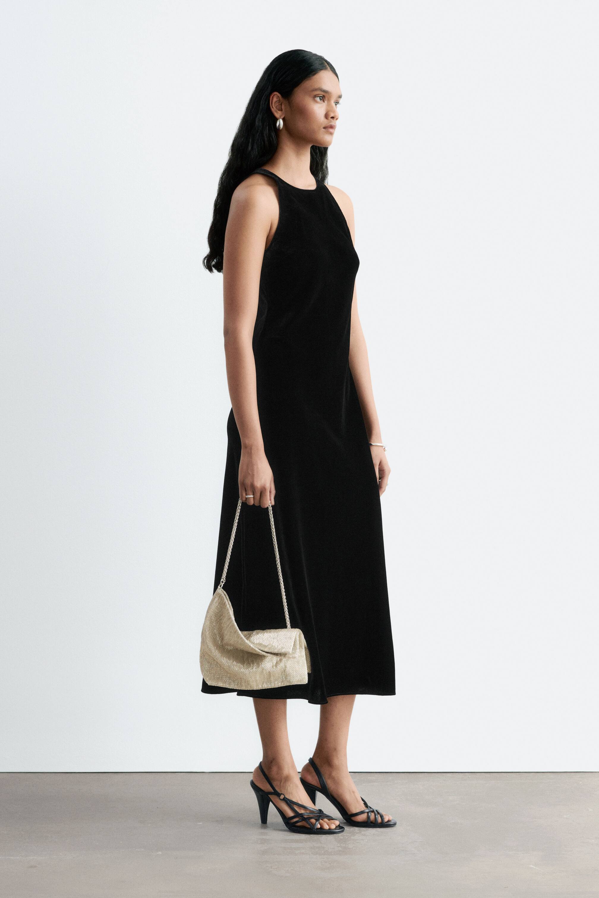 Agrandir l'image: Une robe midi noire sans manches à col rond montant est portée par le mannequin, qui se tient de profil. Le mannequin tient également un sac à bandoulière doré métallisé avec une chaîne et porte des sandales noires à talons hauts à brides.