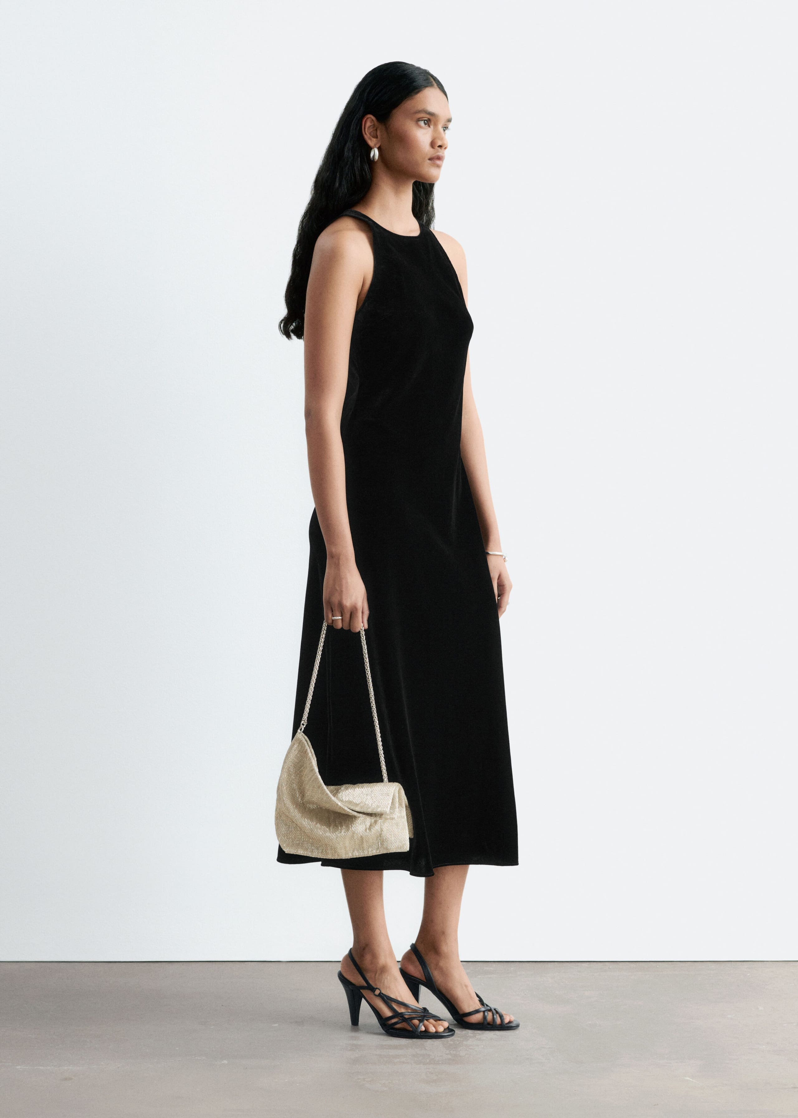A black sleeveless midi-length dress with a high round neckline is being worn by the model, who is standing in profile. The model is also holding a metallic gold shoulder bag with a chain strap and wearing black strappy high-heeled sandals.